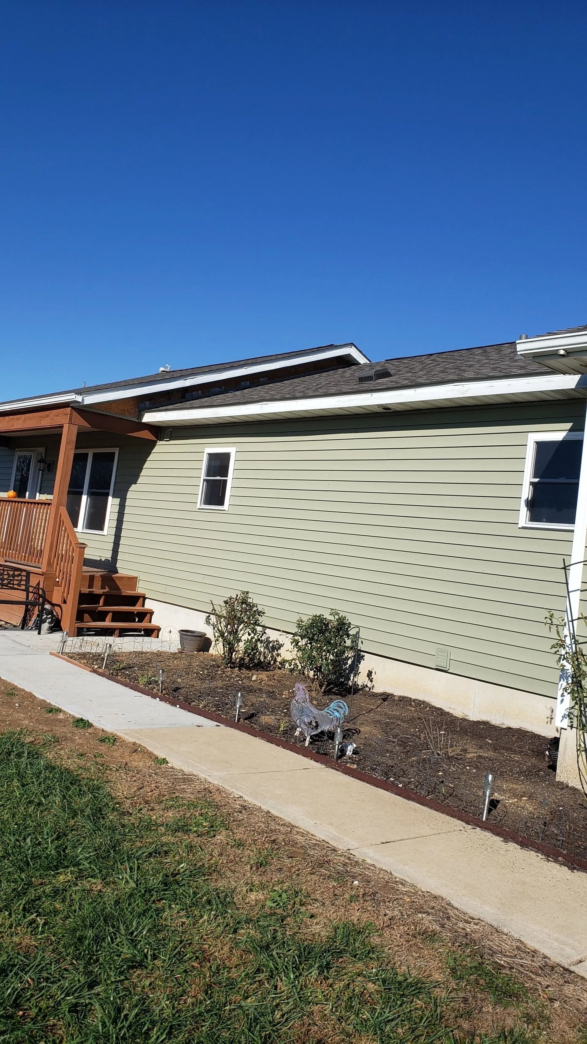 A side view of a green house with a wooden porch, a concrete walkway, and a small garden bed against a clear blue sky.