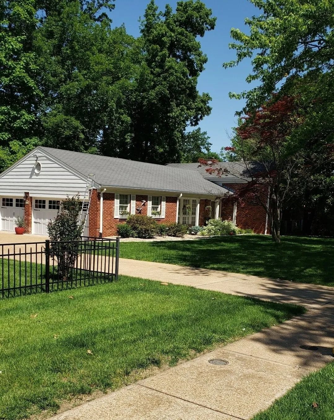 A single-story brick and siding house with a grey shingled roof, set behind a black iron fence in a lush, green yard.