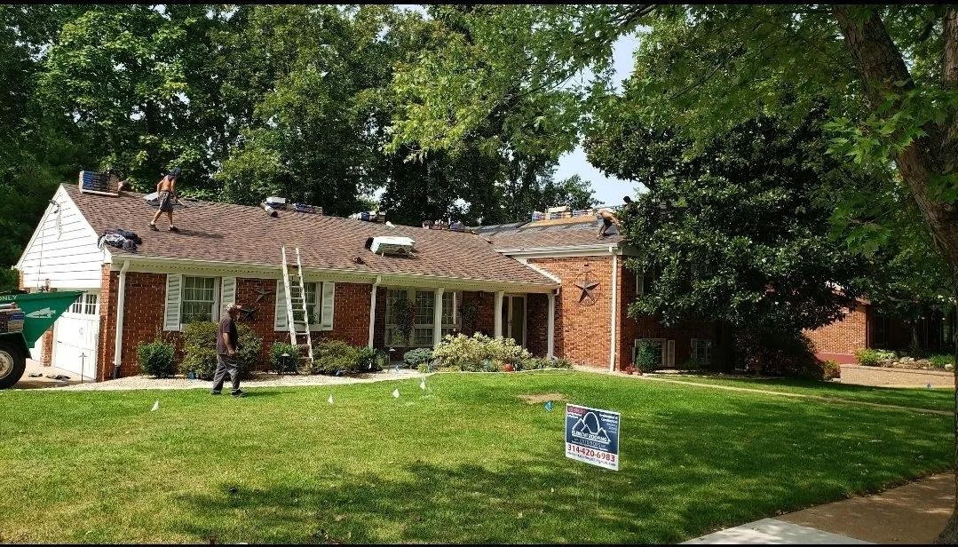 Workers on the roof of a brick house during a residential roofing project, with a green dumpster parked in the yard.