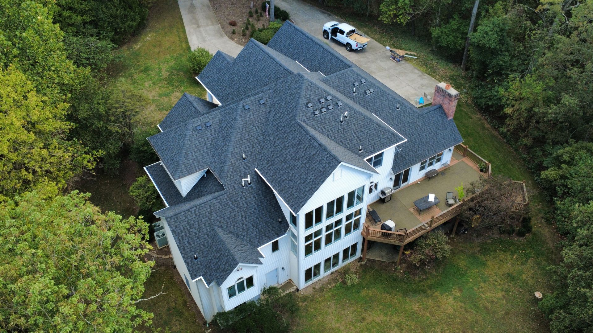 High-angle aerial view of a white multi-story house with a dark gray shingled roof, large deck, and surrounding woods.