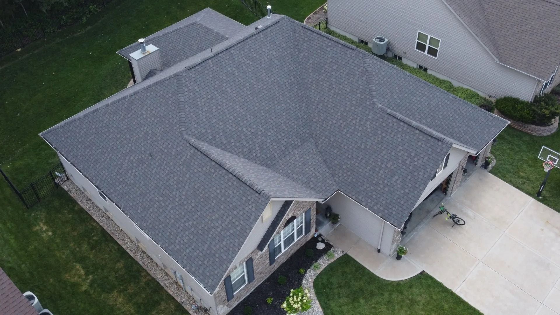 A high-angle aerial view of a residential house with a gray shingled roof, surrounding lawn, and concrete driveway.