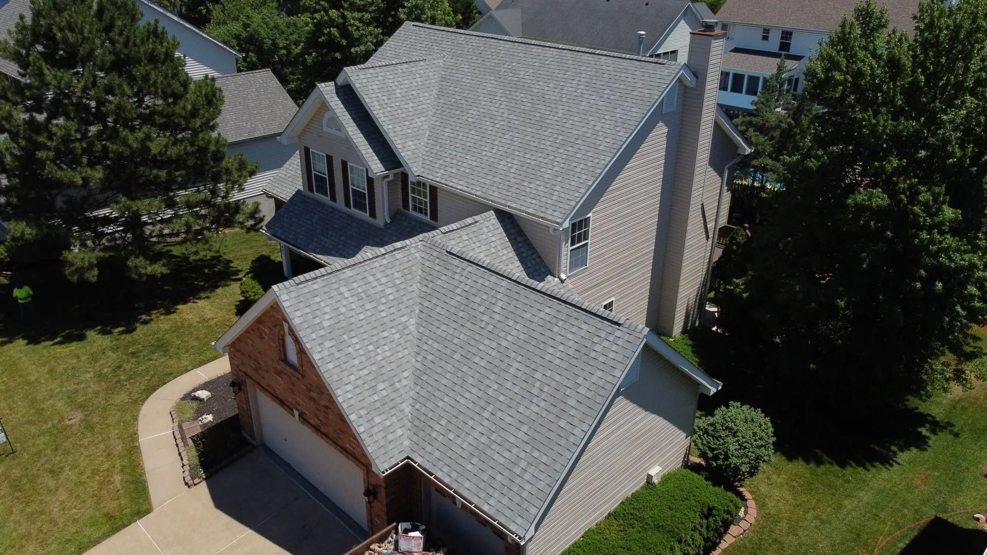 An aerial view of a two-story suburban house with a grey shingled roof, brick accents, and surrounding trees and lawns.