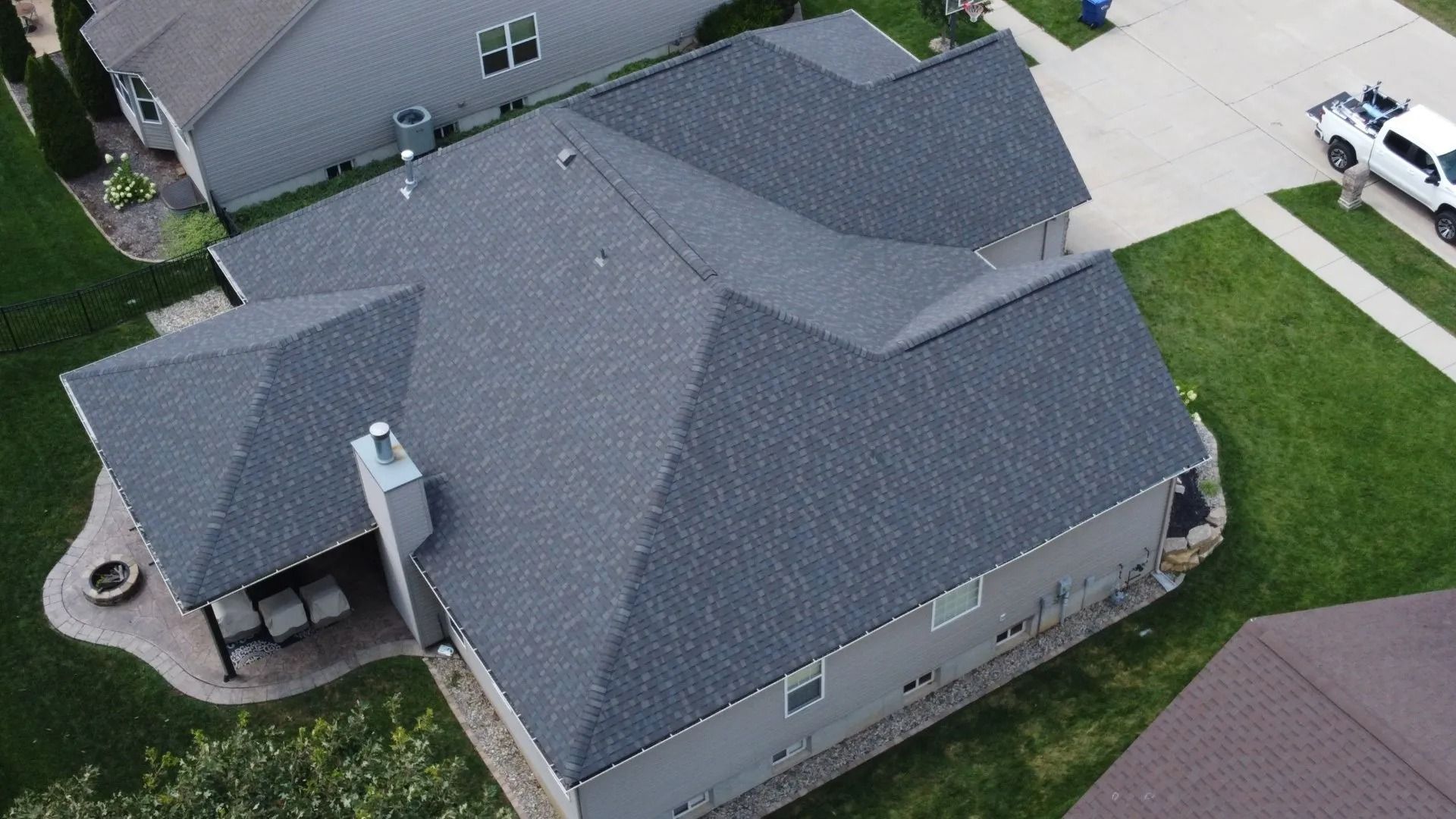 A high-angle aerial view of a gray suburban house with a dark shingled roof, a chimney, and a backyard patio.