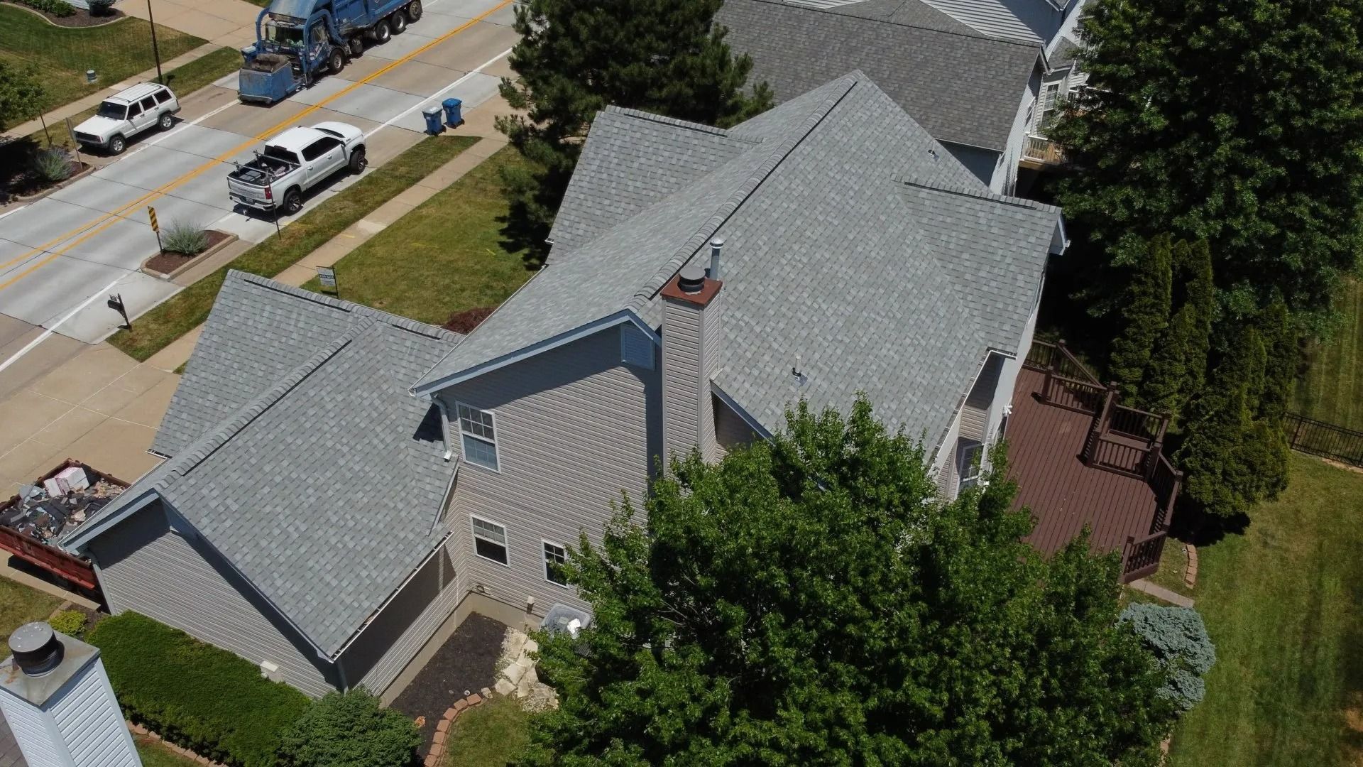 An aerial view of a light-colored house with a grey shingled roof, surrounded by trees, a deck, and a nearby street.