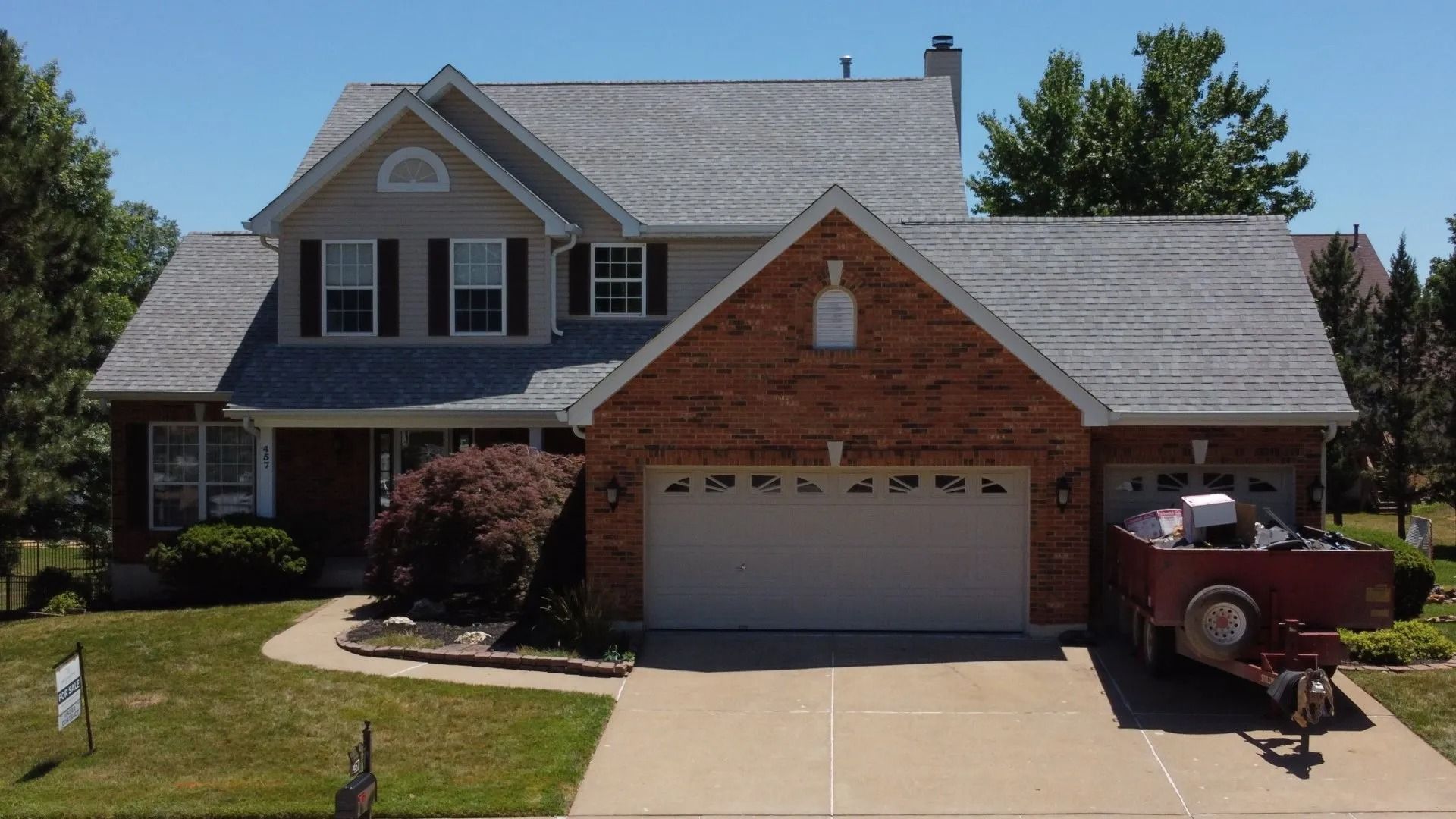 A two-story suburban house with a brick front, grey roof, attached garage, and a trailer parked on the driveway.
