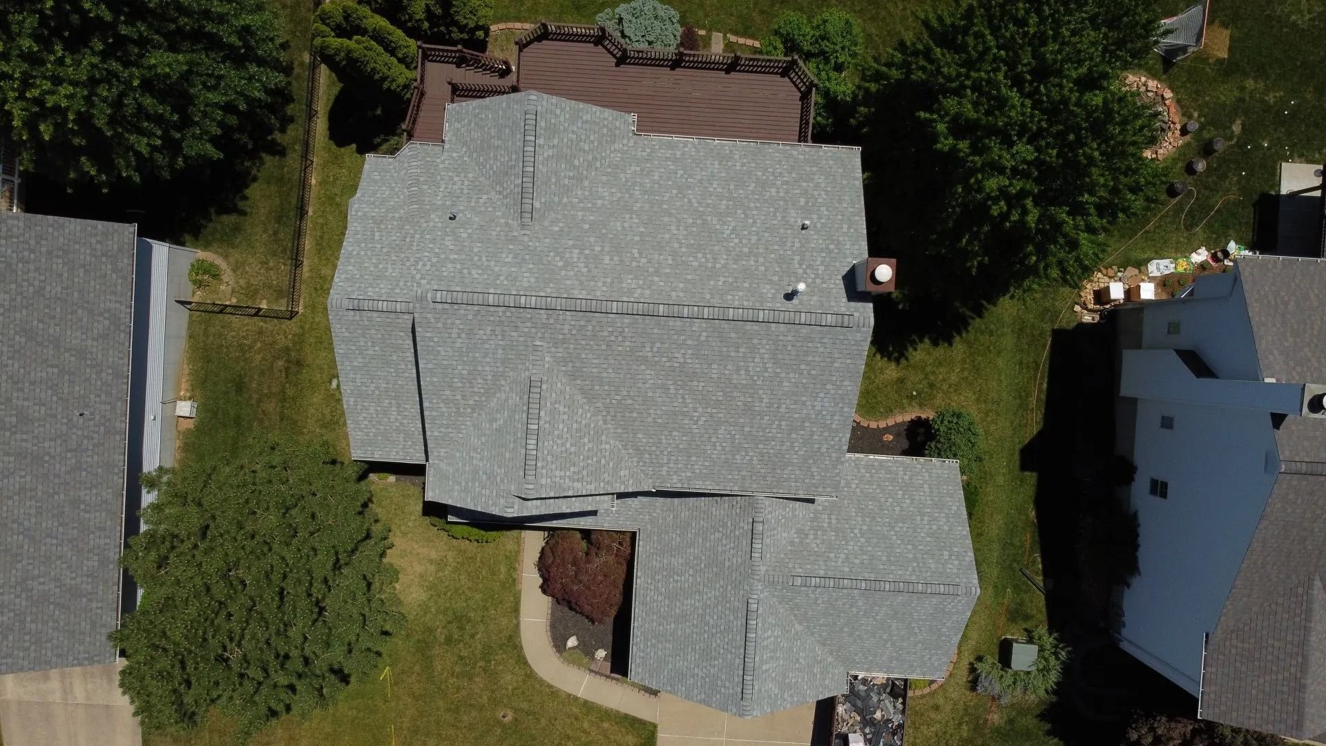 An aerial top-down view of a house with a gray shingled roof, surrounded by green grass and trees.