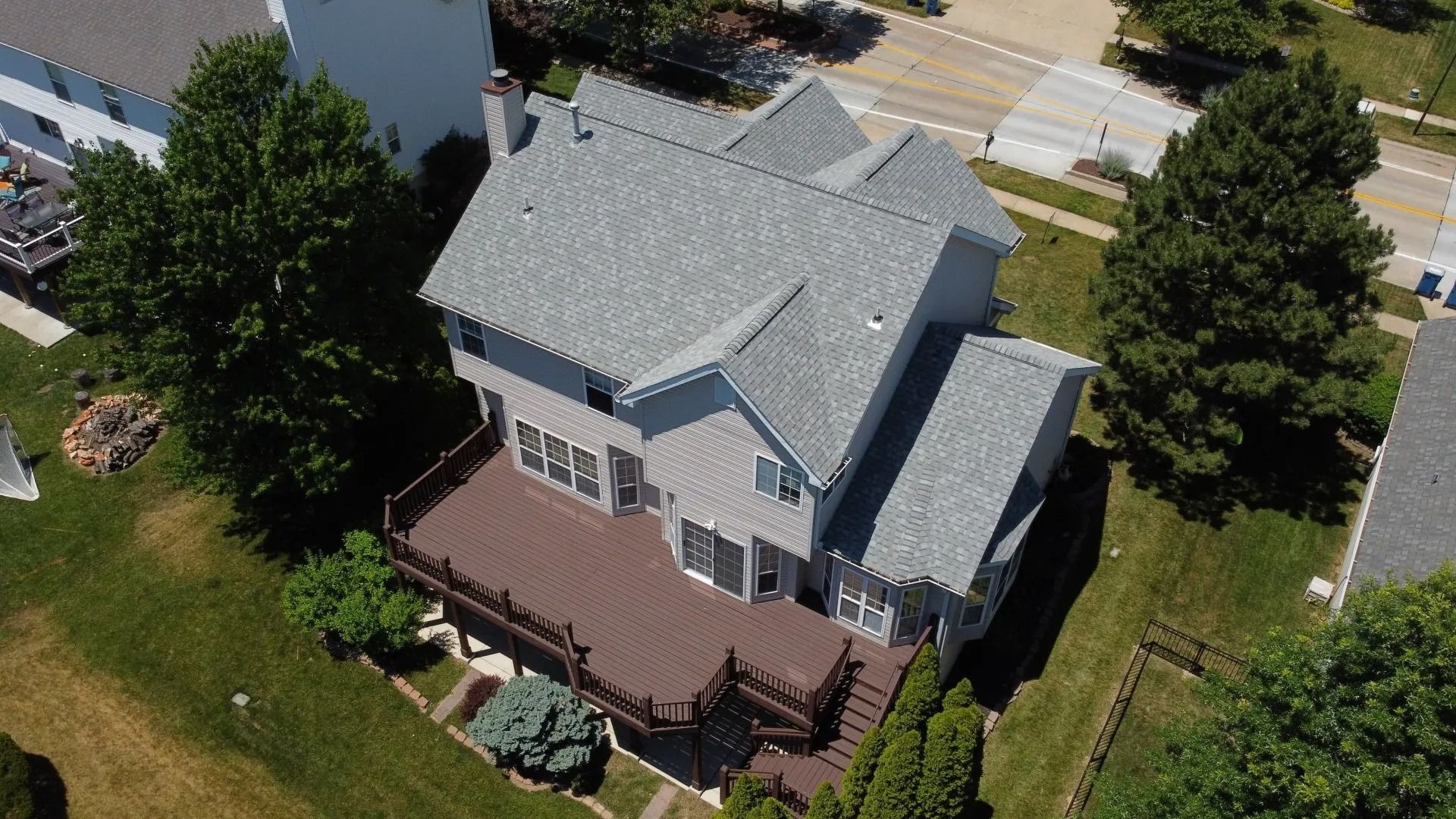 An aerial view of a two-story gray house with a large brown deck and a shingled roof, surrounded by trees and lawn.