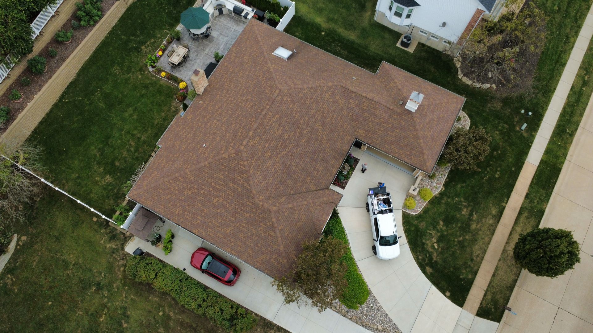 An aerial view of a single-story house with a brown shingled roof, a paved driveway with two cars, and a backyard patio.
