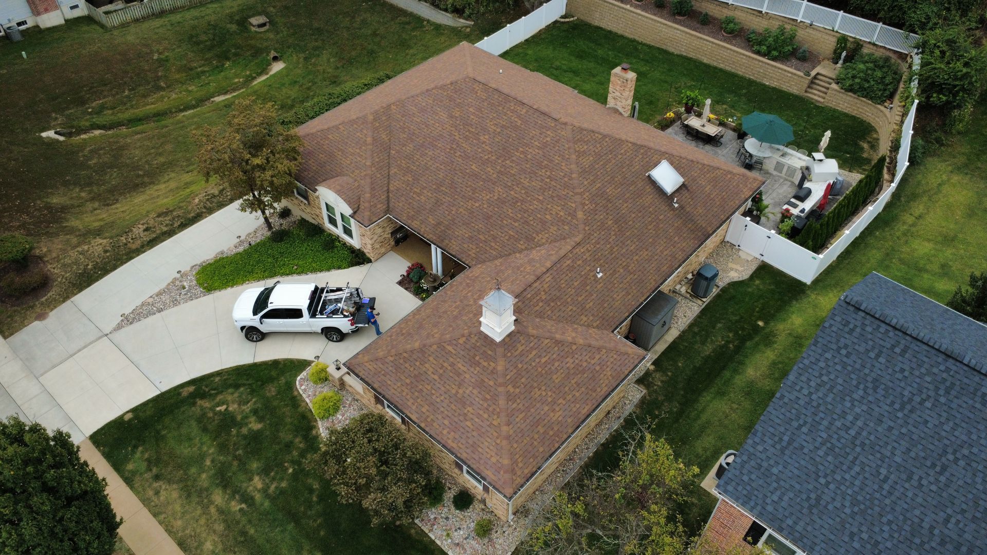An aerial view of a tan-brick residential house with a brown shingled roof, a driveway with a truck, and a backyard.
