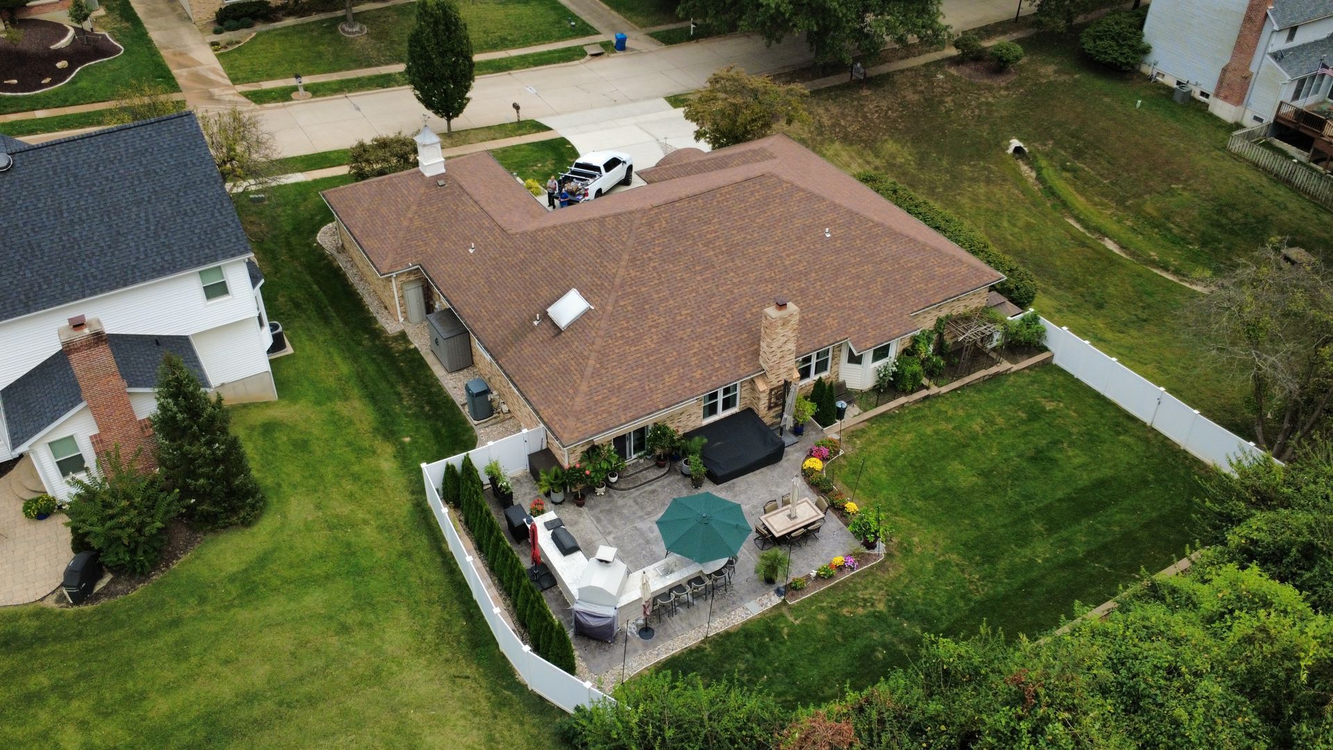 An aerial view of a suburban house with a brown shingled roof, a stone patio with a green umbrella, and a grassy backyard.