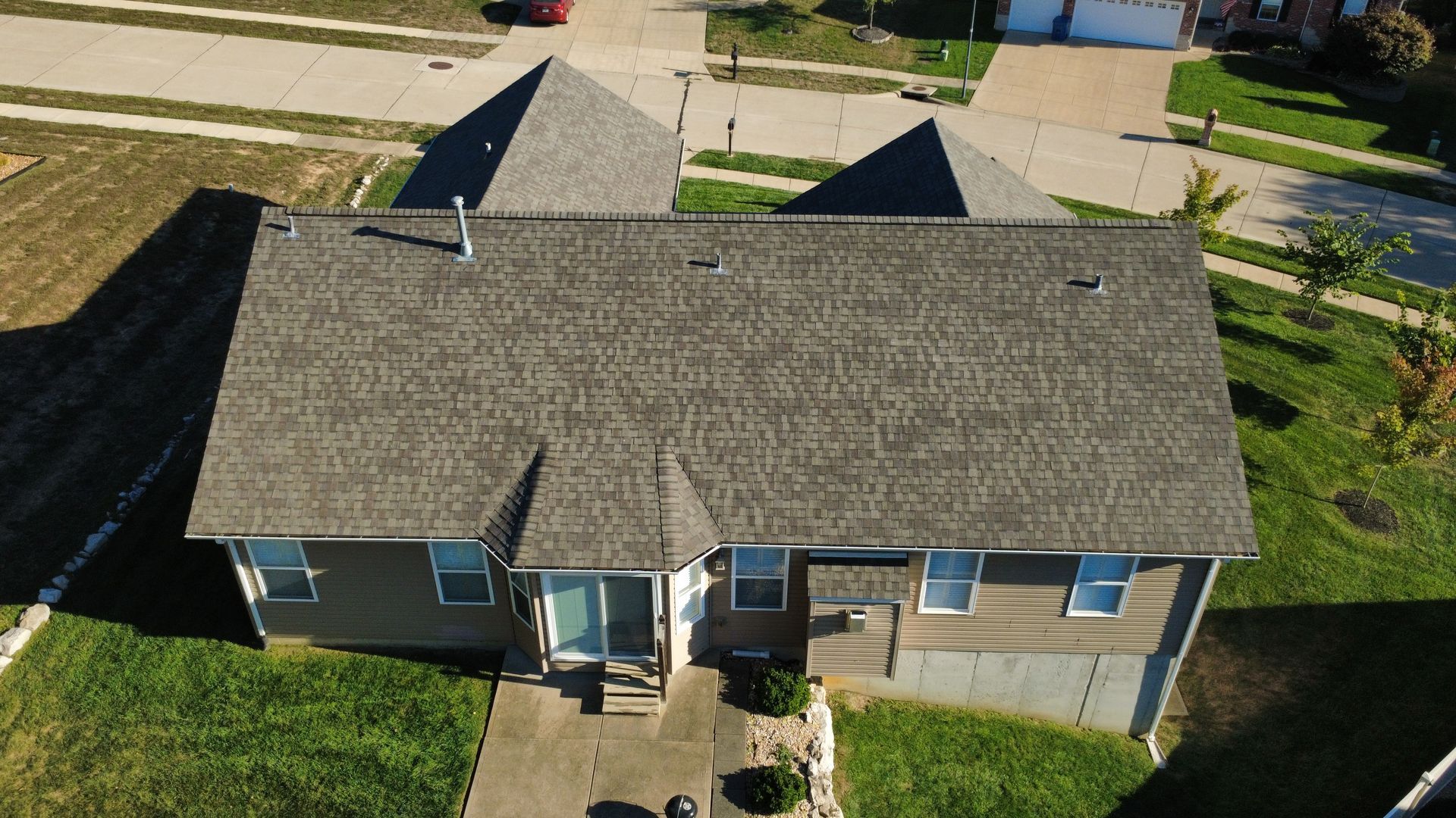 Aerial view of a single-story home with a gray shingled roof, brown siding, and a concrete driveway in a suburban area.