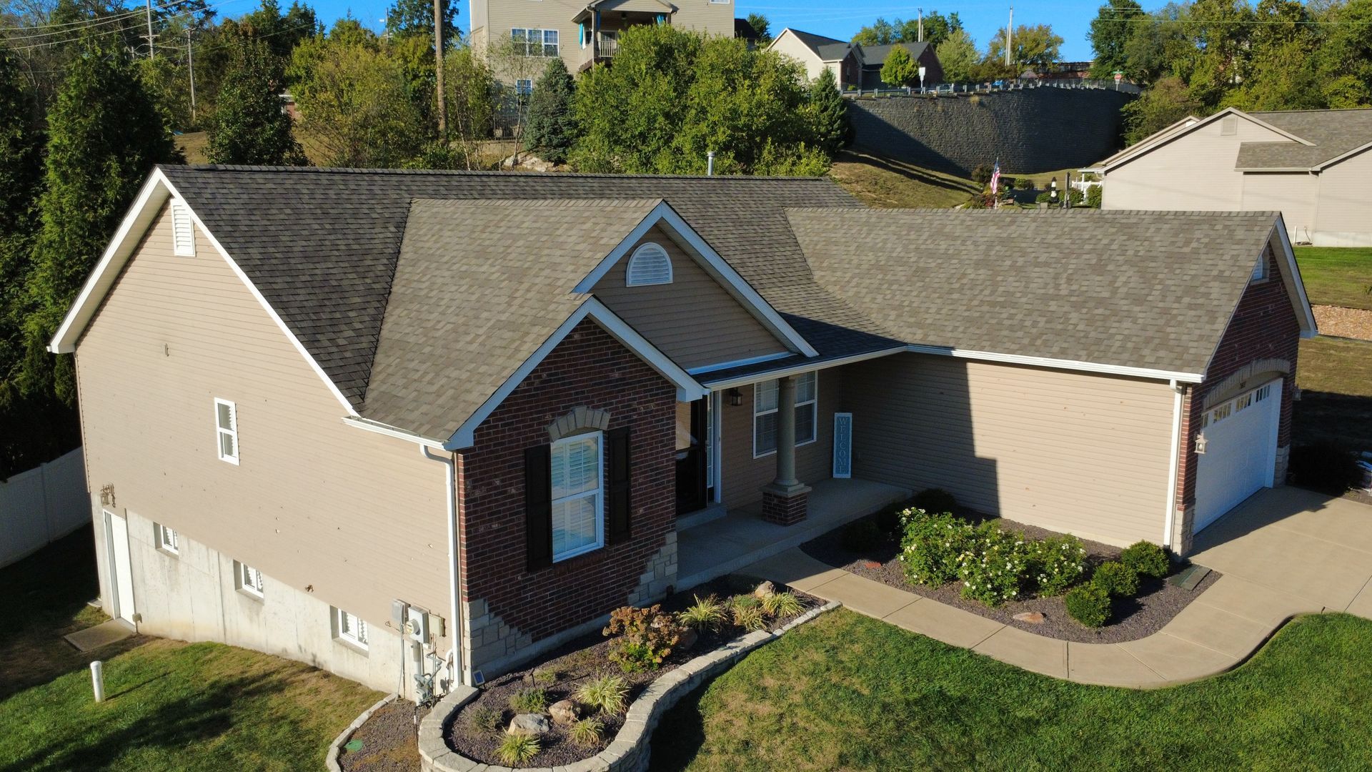 An aerial view of a tan, single-story suburban home with a brick entryway, gray roof, and attached garage.