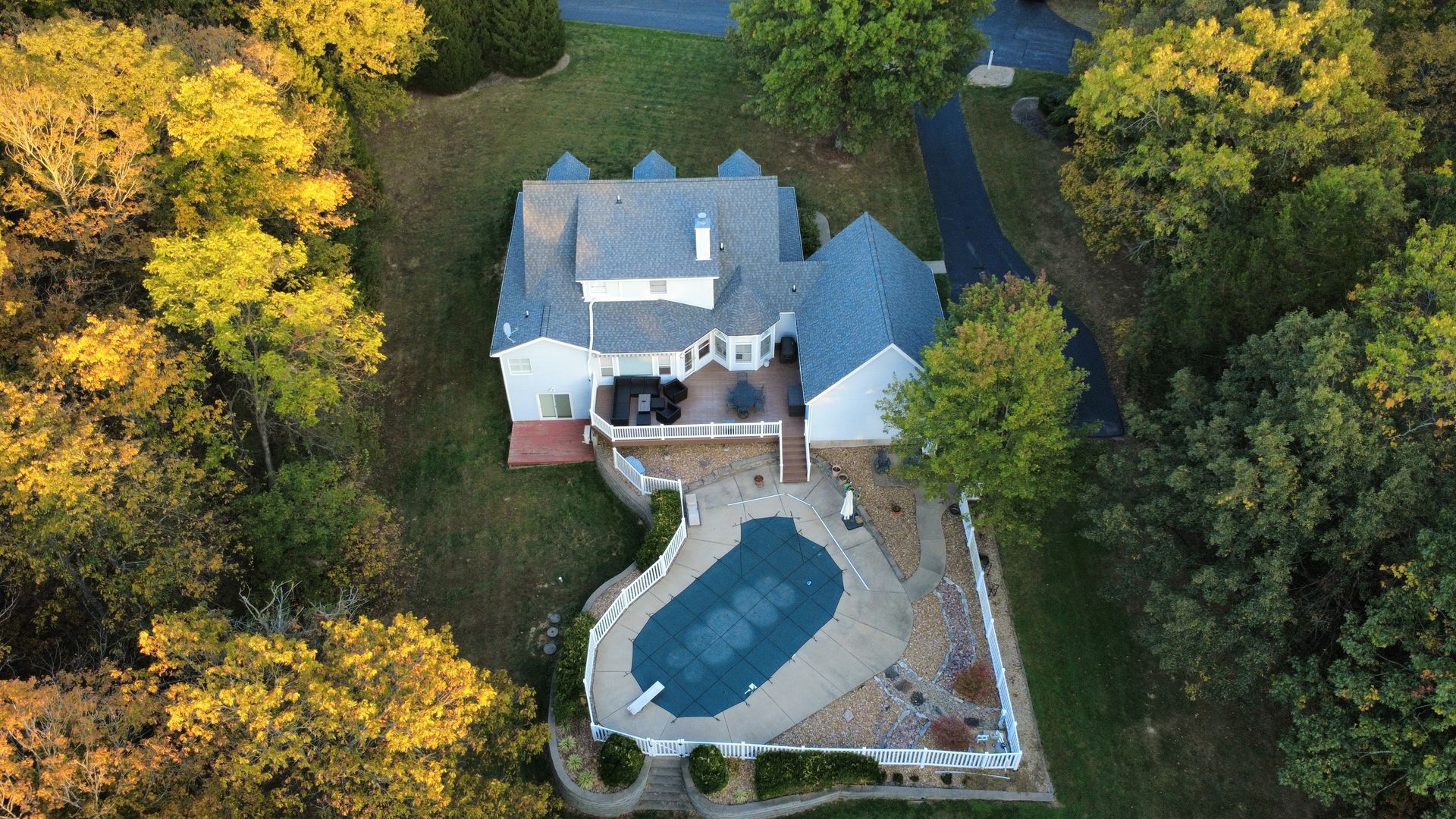 An aerial view of a two-story home with a swimming pool, surrounded by wooded grounds in autumn.