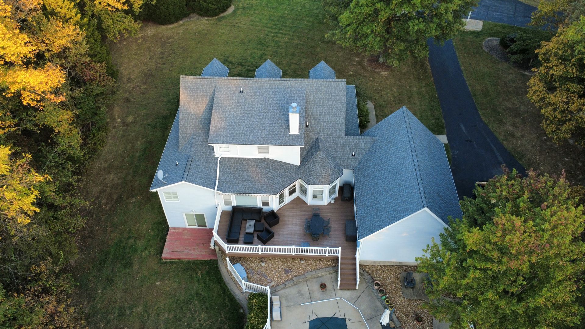 Aerial view of a house with a gray roof, a deck, and a patio, surrounded by trees and a driveway.