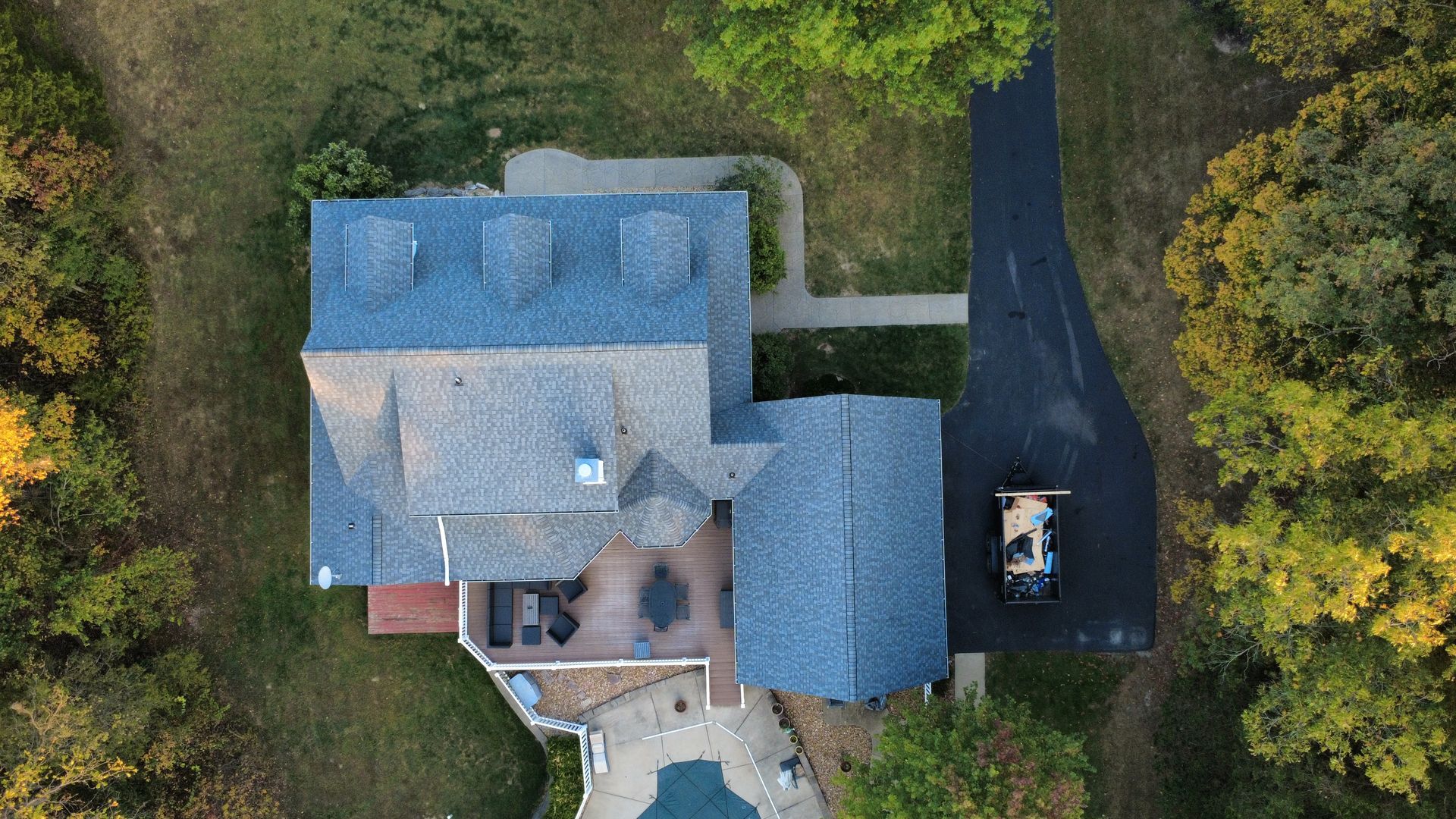 An aerial view of a house with a gray shingled roof, a paved driveway, a backyard deck, and a pool, surrounded by trees.