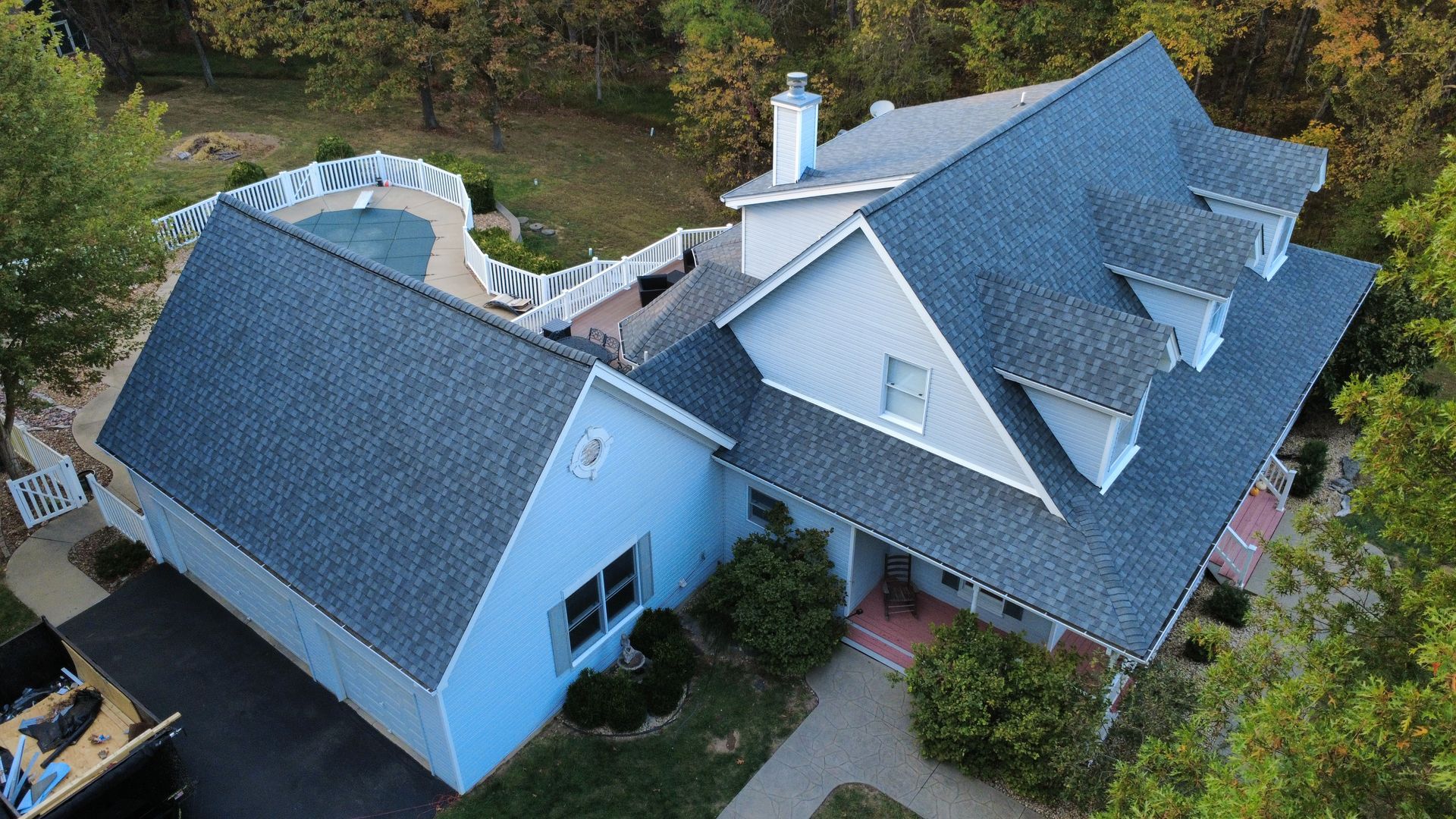 An aerial view of a light blue house with a gray shingled roof, a swimming pool in the yard, and surrounding trees.