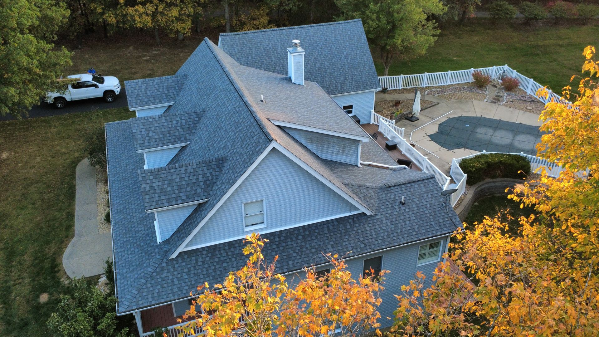 An aerial view of a blue-sided house with a dark shingled roof, a chimney, and a fenced-in swimming pool in the backyard.