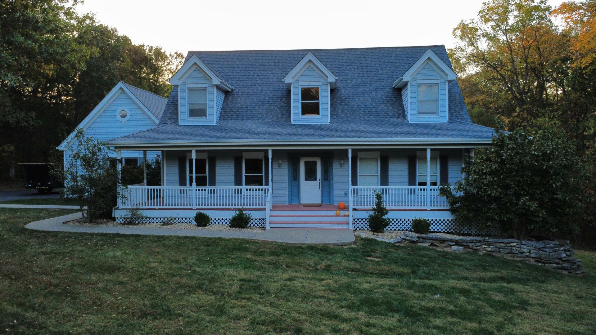A two-story light blue house with a wide front porch, three dormer windows, and a paved walkway, set among trees.