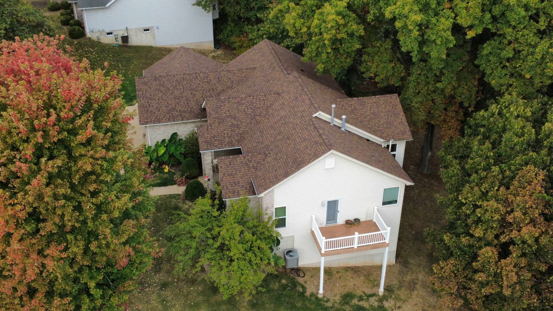 Aerial view of a white house with a brown shingled roof, surrounded by trees, with a small raised deck in the back.