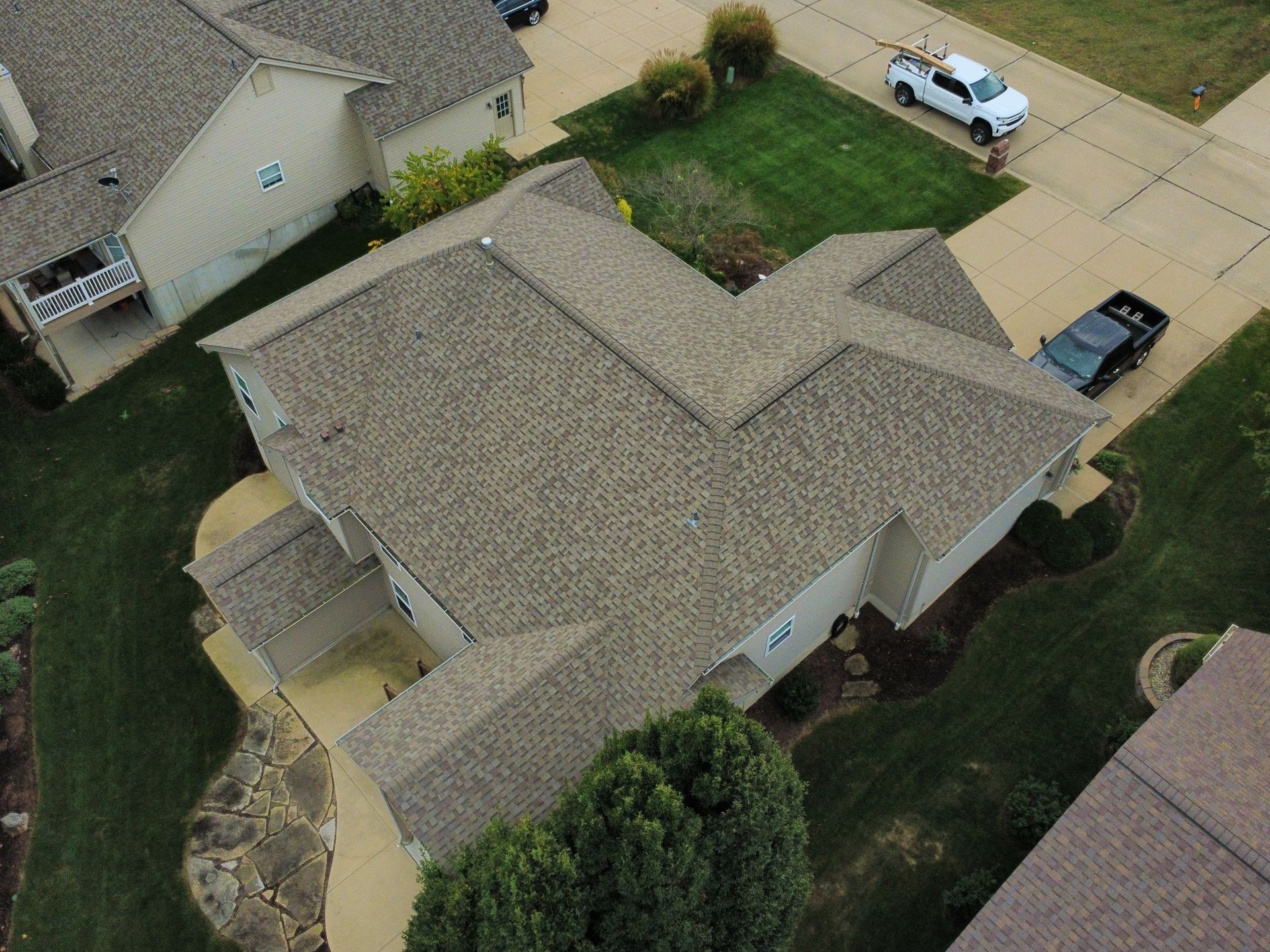An aerial view of a tan-colored residential home with a shingled roof, surrounded by a lawn and concrete driveway.