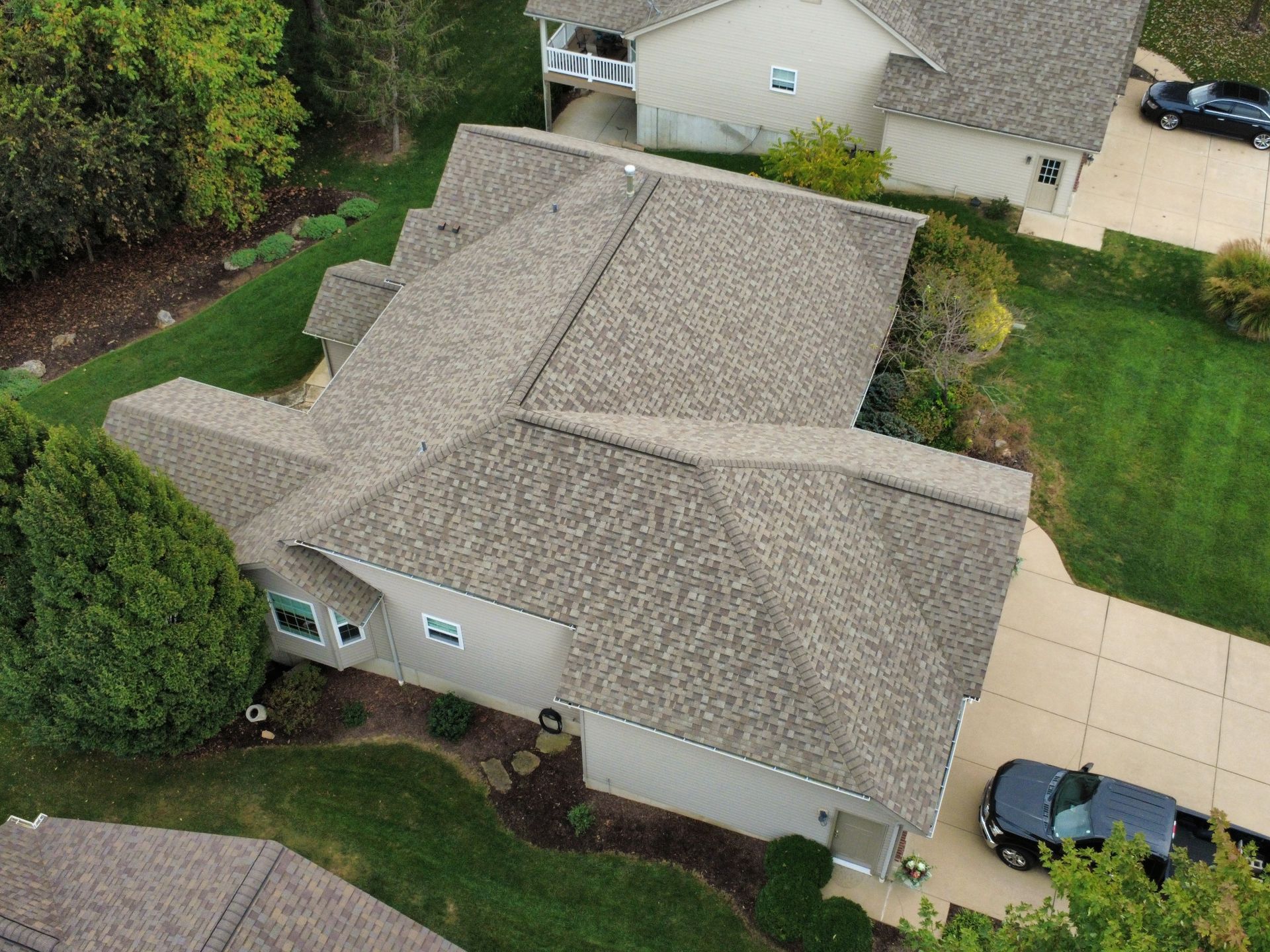 Aerial view of a residential home with a tan shingled roof, surrounded by green lawns, trees, and paved driveways.