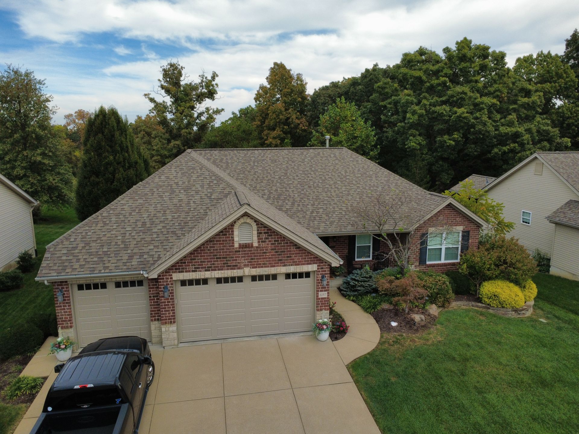 An aerial view of a brick house with a tan garage and a shingled roof, set in a suburban neighborhood with green trees.