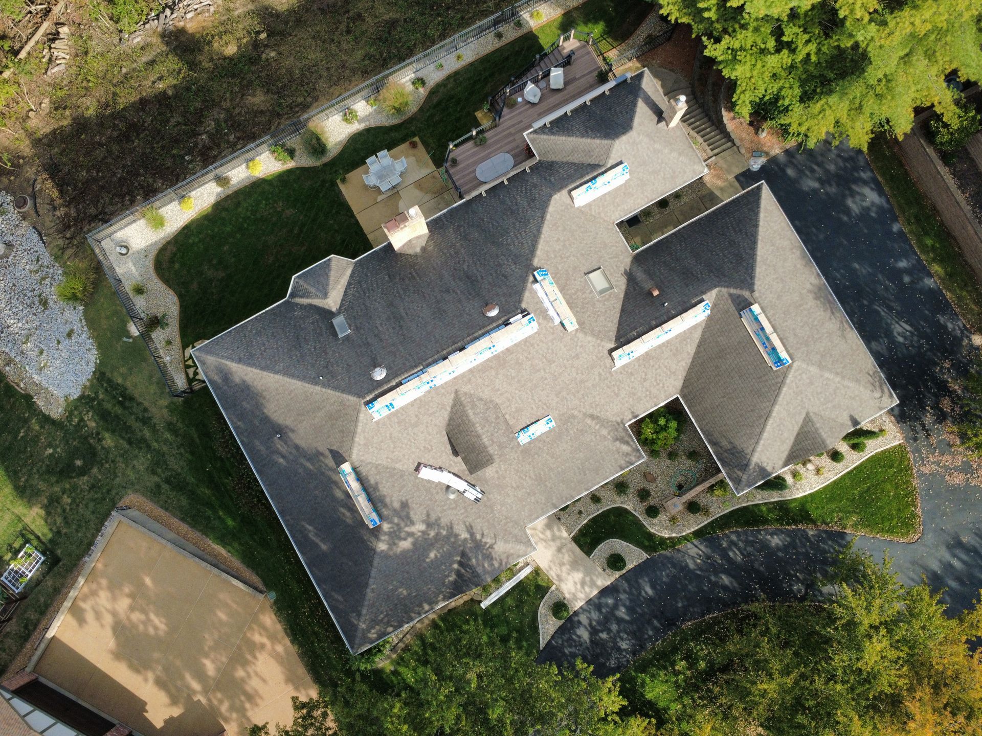 A top-down aerial view of a large residential house with a dark shingled roof, surrounded by a lawn, trees, and driveway.