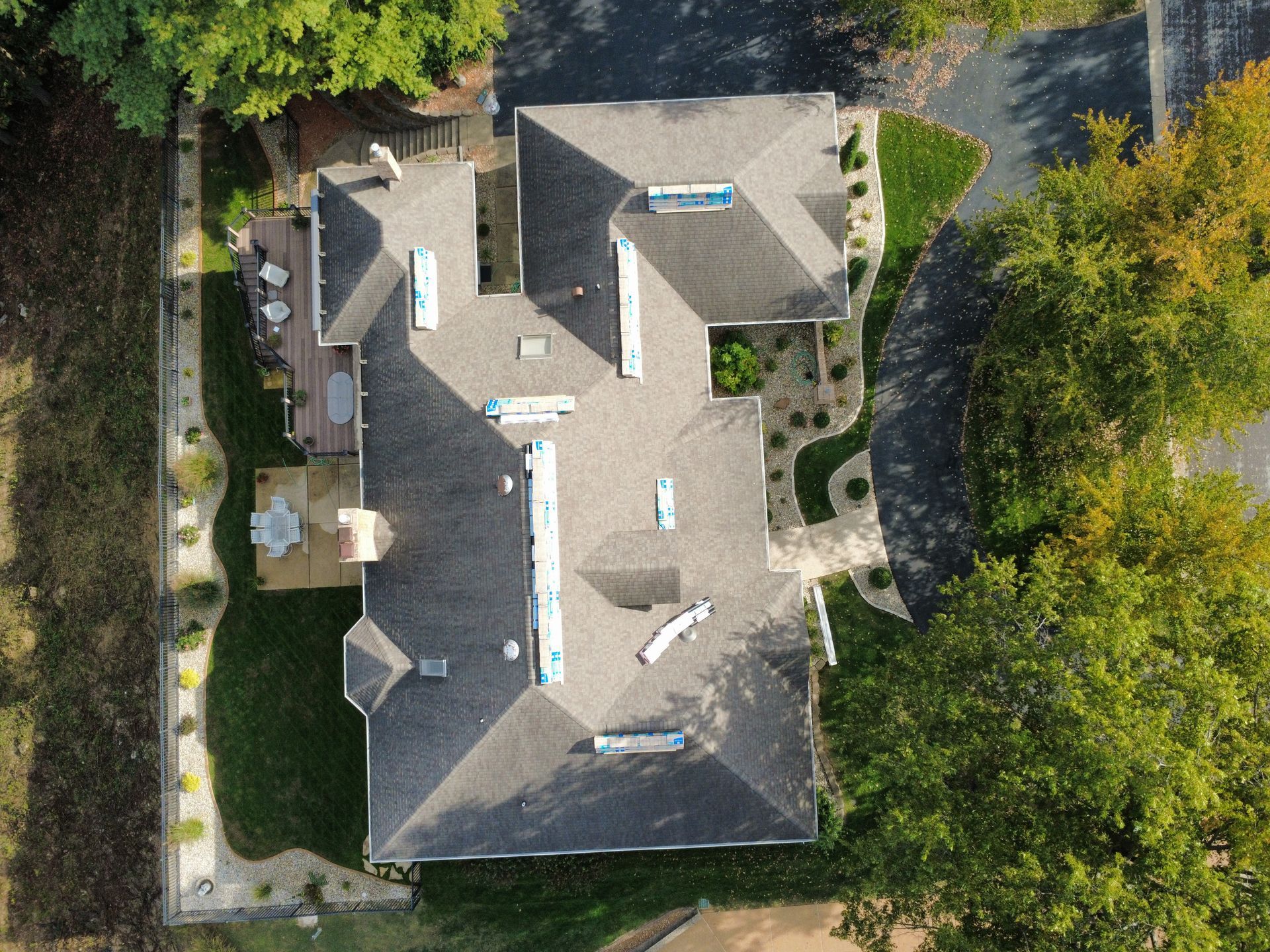 A direct overhead drone shot of a multi-gabled house with a grey shingled roof, surrounded by trees and a paved driveway.