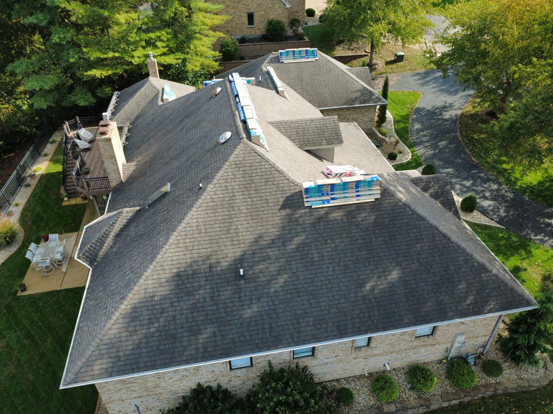 High-angle aerial view of a brick house with a dark shingled roof under repair, featuring bundles of tiles on top.