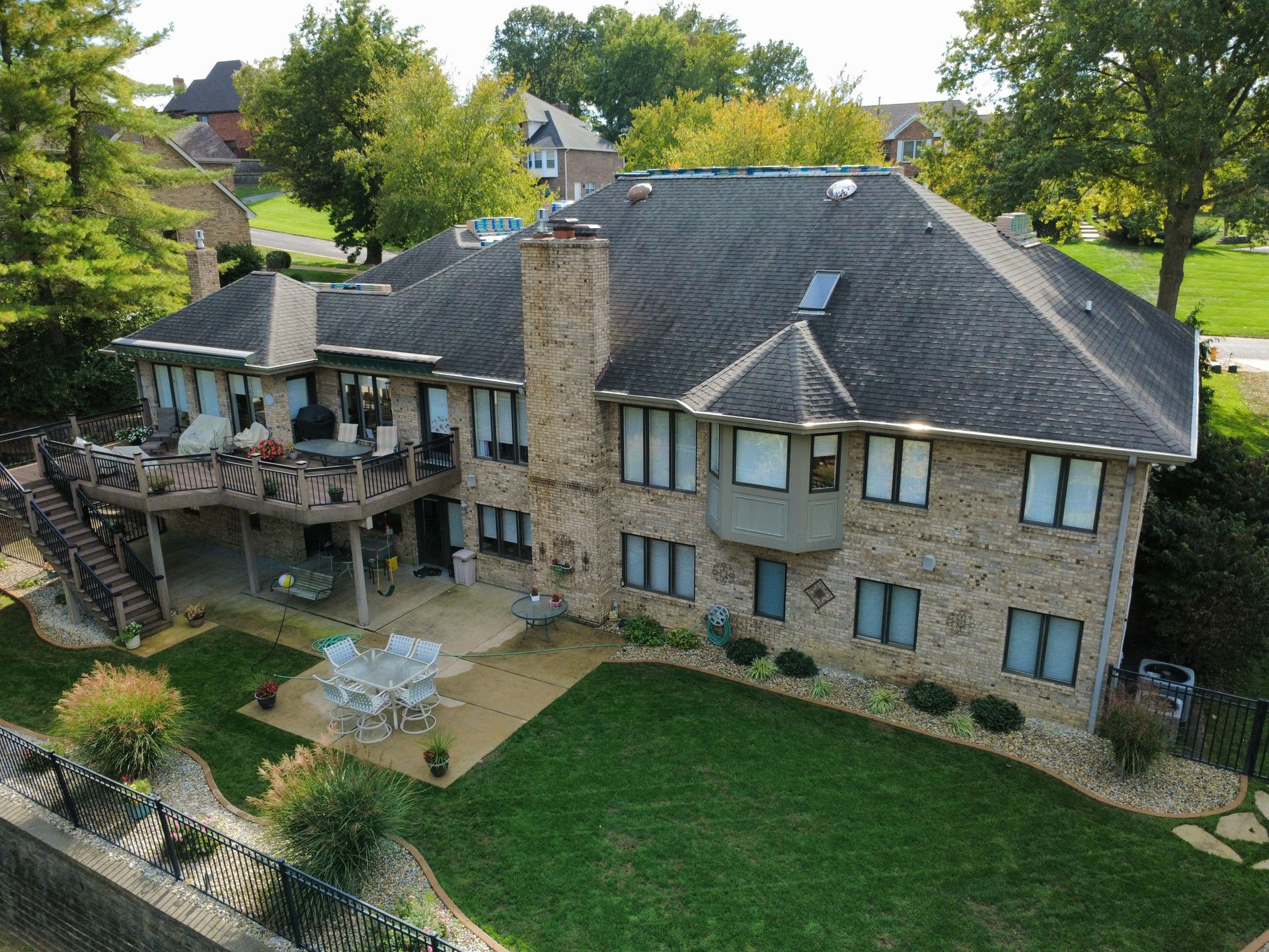 A multi-story stone house with a large wooden deck, patio, and backyard surrounded by trees on a sunny day.