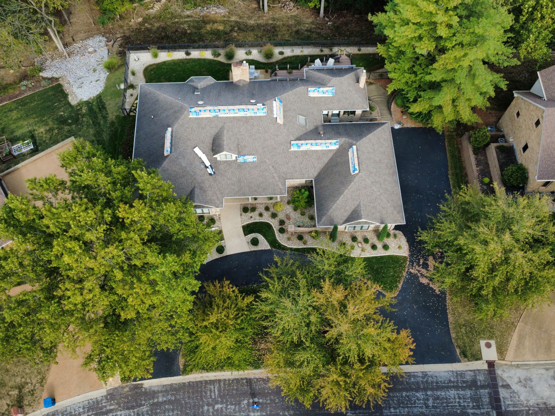 Aerial view of a house with gray roofing shingles surrounded by trees and a paved driveway.