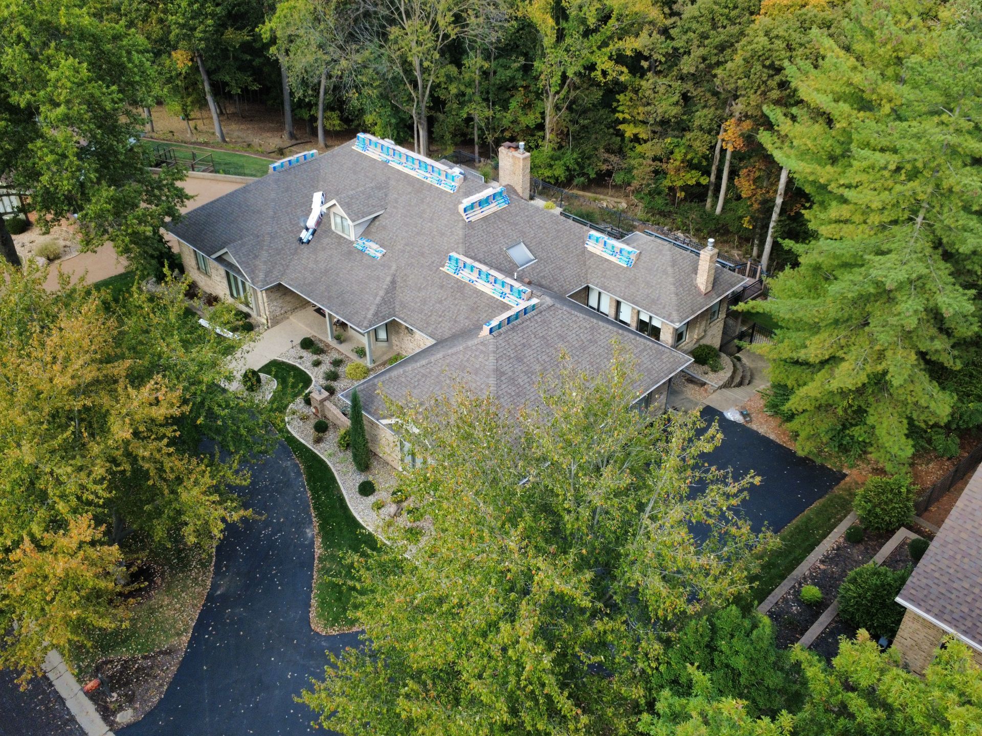 An aerial view of a suburban house with a gray roof, surrounded by trees and a paved driveway.