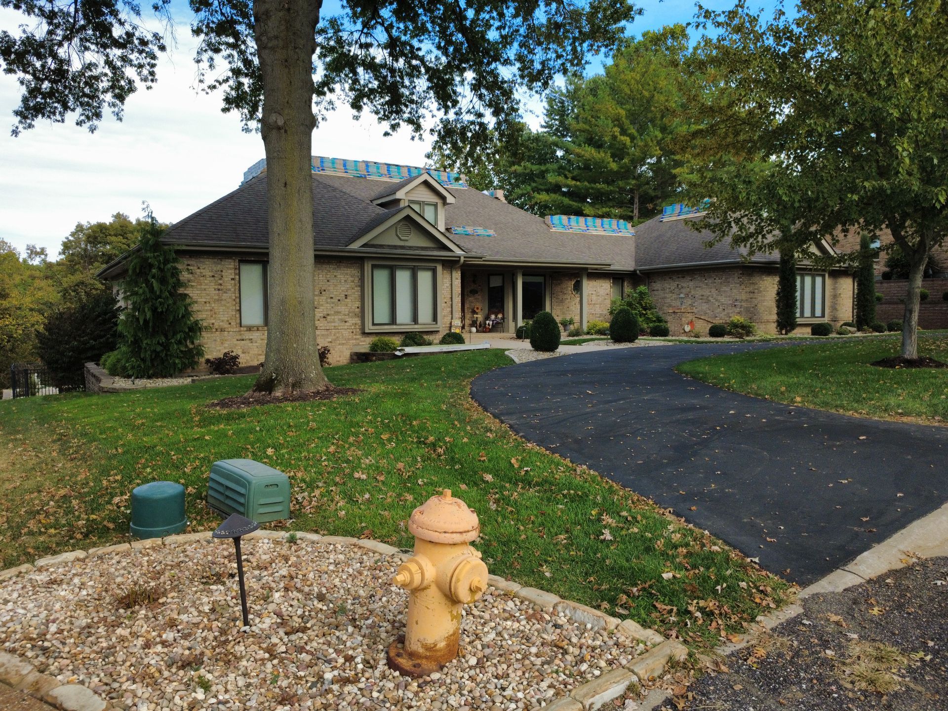 A tan brick house with a dark shingled roof, a curved asphalt driveway, and a yellow fire hydrant in the foreground.