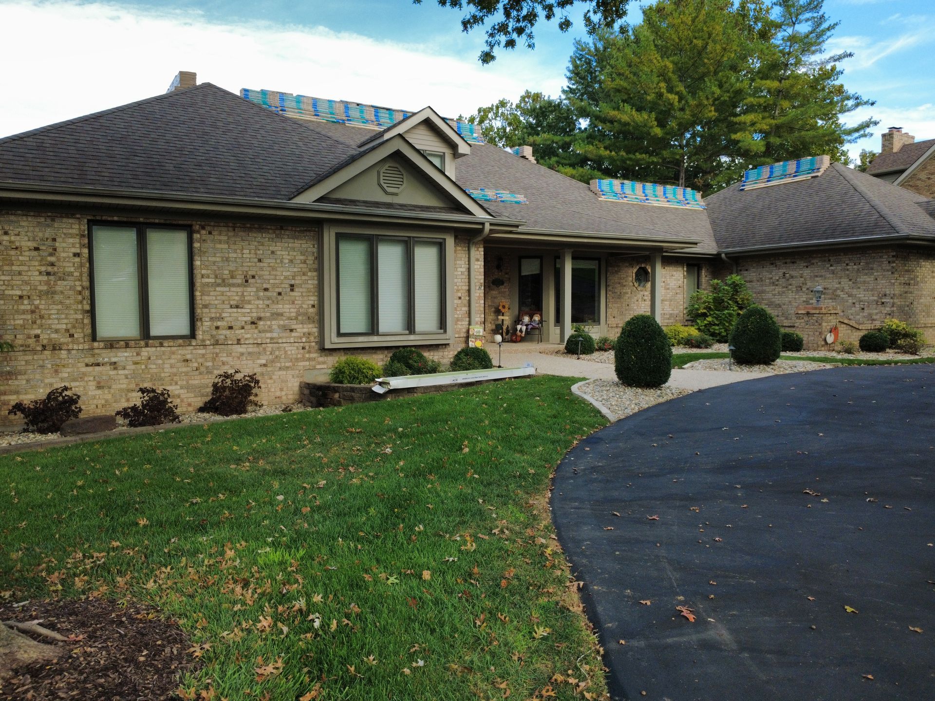A beige brick house with a dark roof under repair, featuring a front lawn and a curved asphalt driveway.