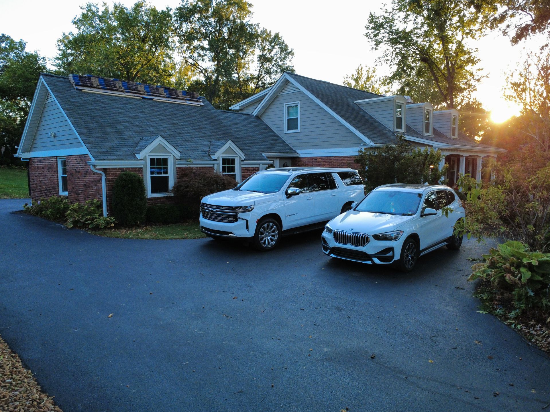 A white SUV and a white sedan are parked on a paved driveway in front of a brick house with gray roof shingles at sunset.
