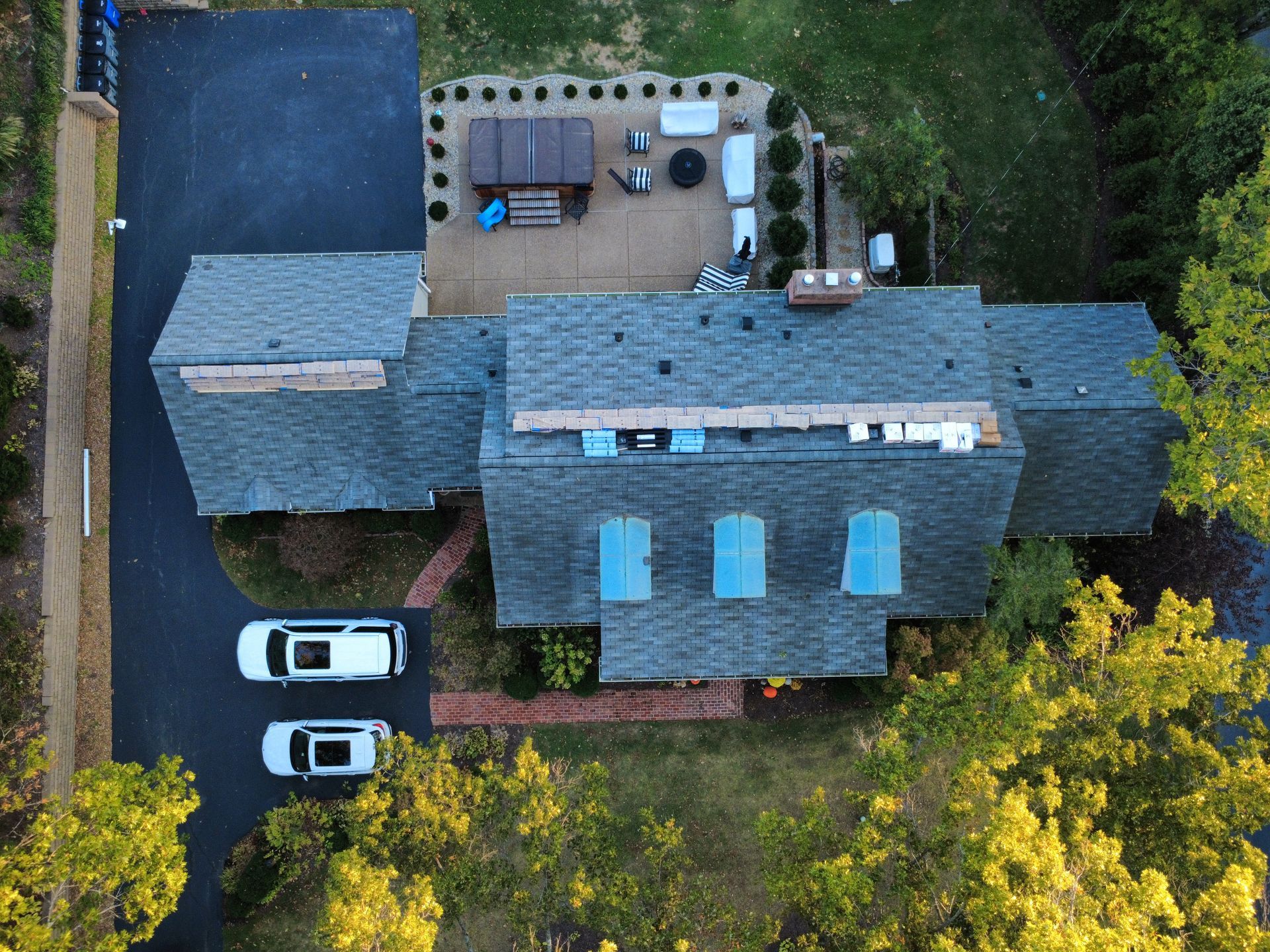 Aerial view of a residential house with a shingled roof, a back patio with a hot tub, and two white cars in the driveway.