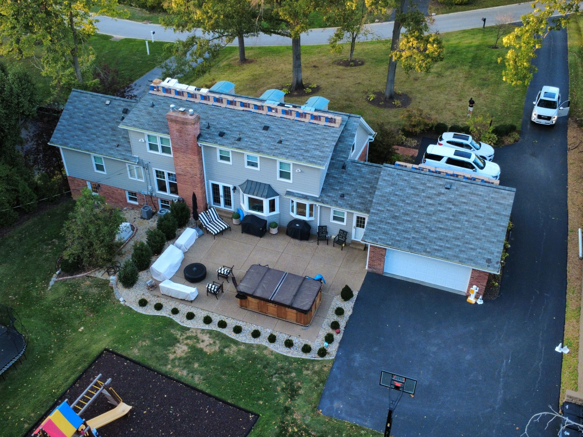 Aerial view of a two-story gray house with a stone patio, hot tub, large driveway, and backyard playground.