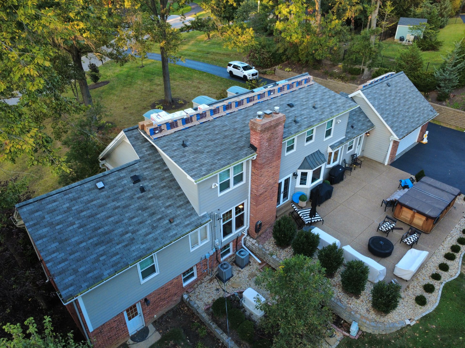 Aerial view of a two-story house with a grey shingled roof, brick chimney, and a rear patio with outdoor furniture.