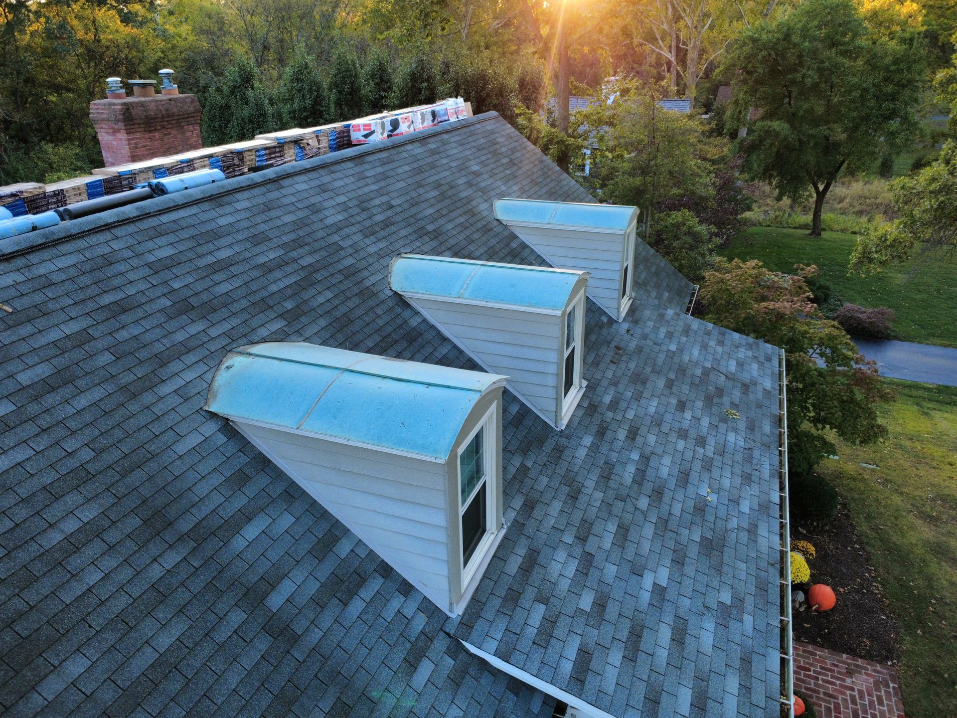 An aerial view of a dark shingled roof with three dormer windows and bundles of new roofing shingles placed at the ridge.