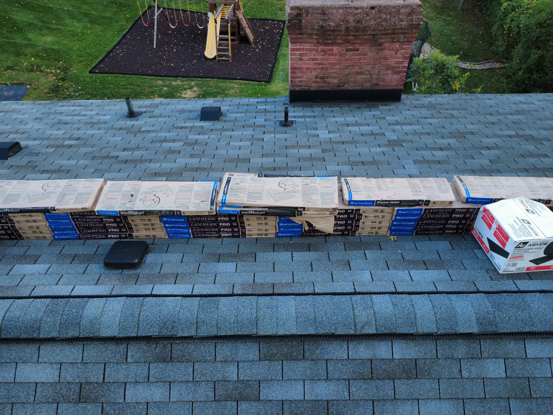 Several stacks of roofing shingles arranged in a long row on a gray shingled roof next to a brick chimney.