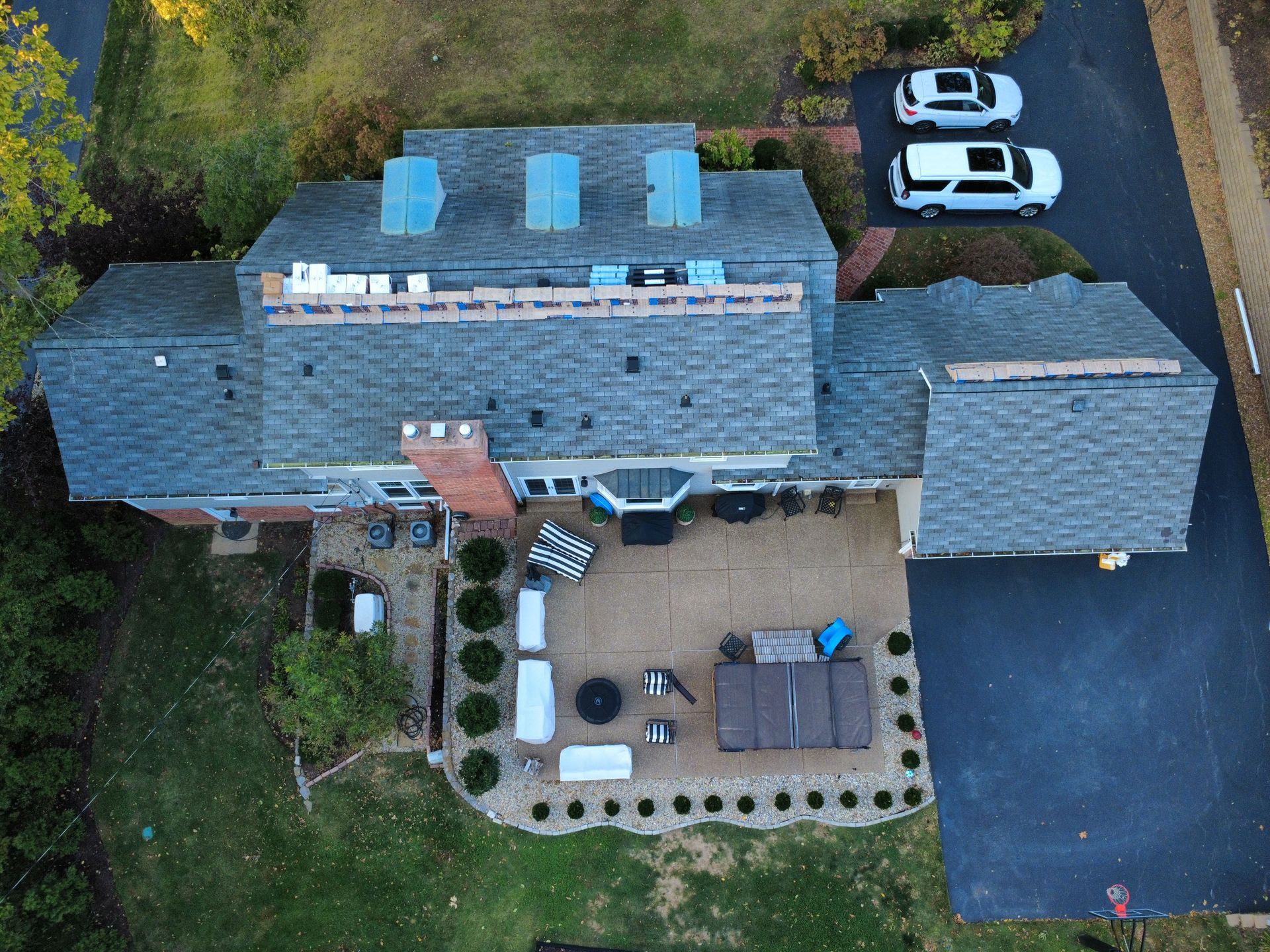 Aerial view of a residential house with a shingled roof under repair, a patio area with furniture, and a driveway with cars.
