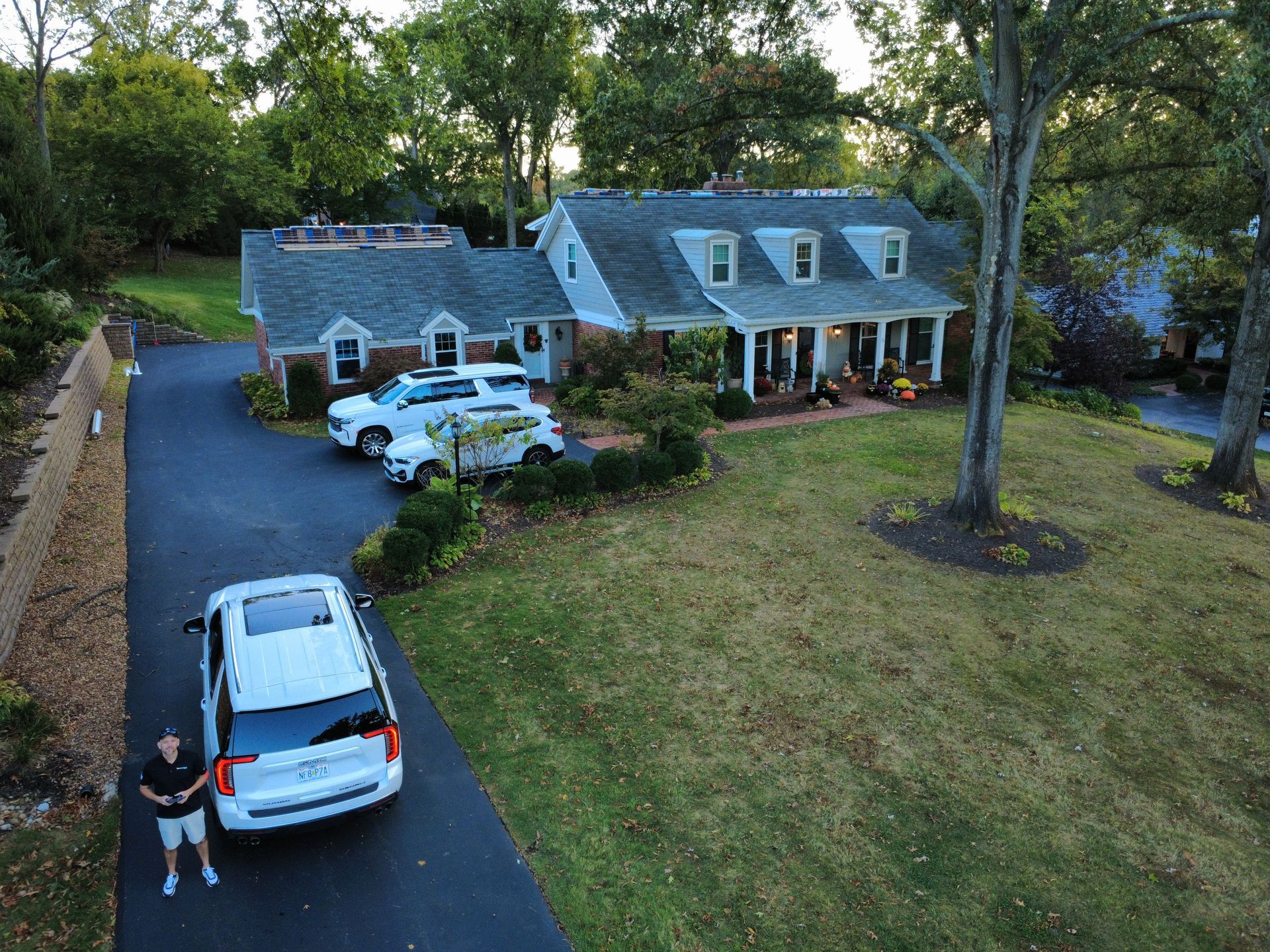 Aerial view of a house with a driveway, three parked white SUVs, and a person standing near one of the vehicles.