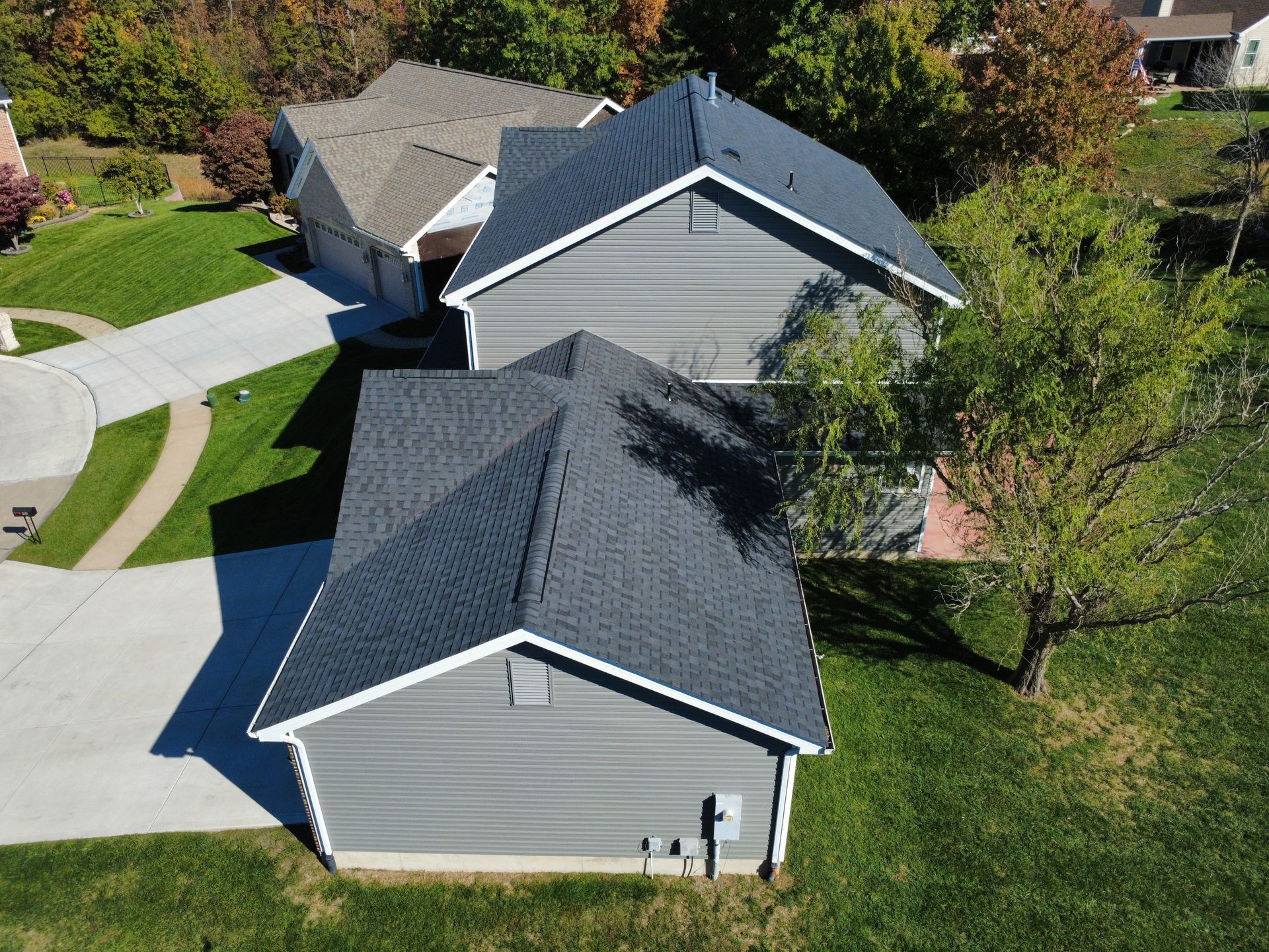 An aerial view of two residential houses with gray siding and dark shingled roofs surrounded by green lawns and trees.
