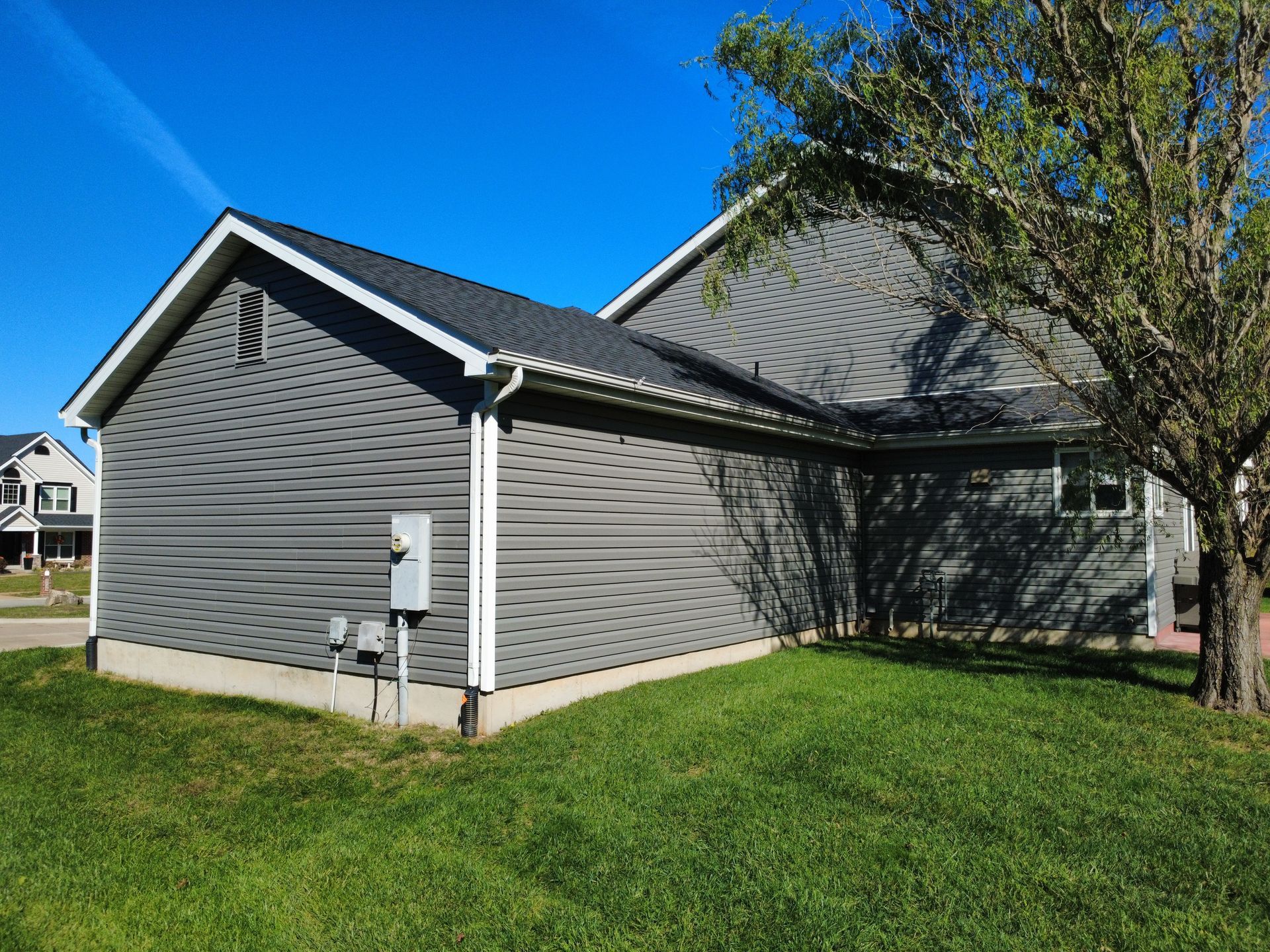 A gray-sided house with a dark metal roof and white trim sits on a grassy lawn under a clear blue sky.