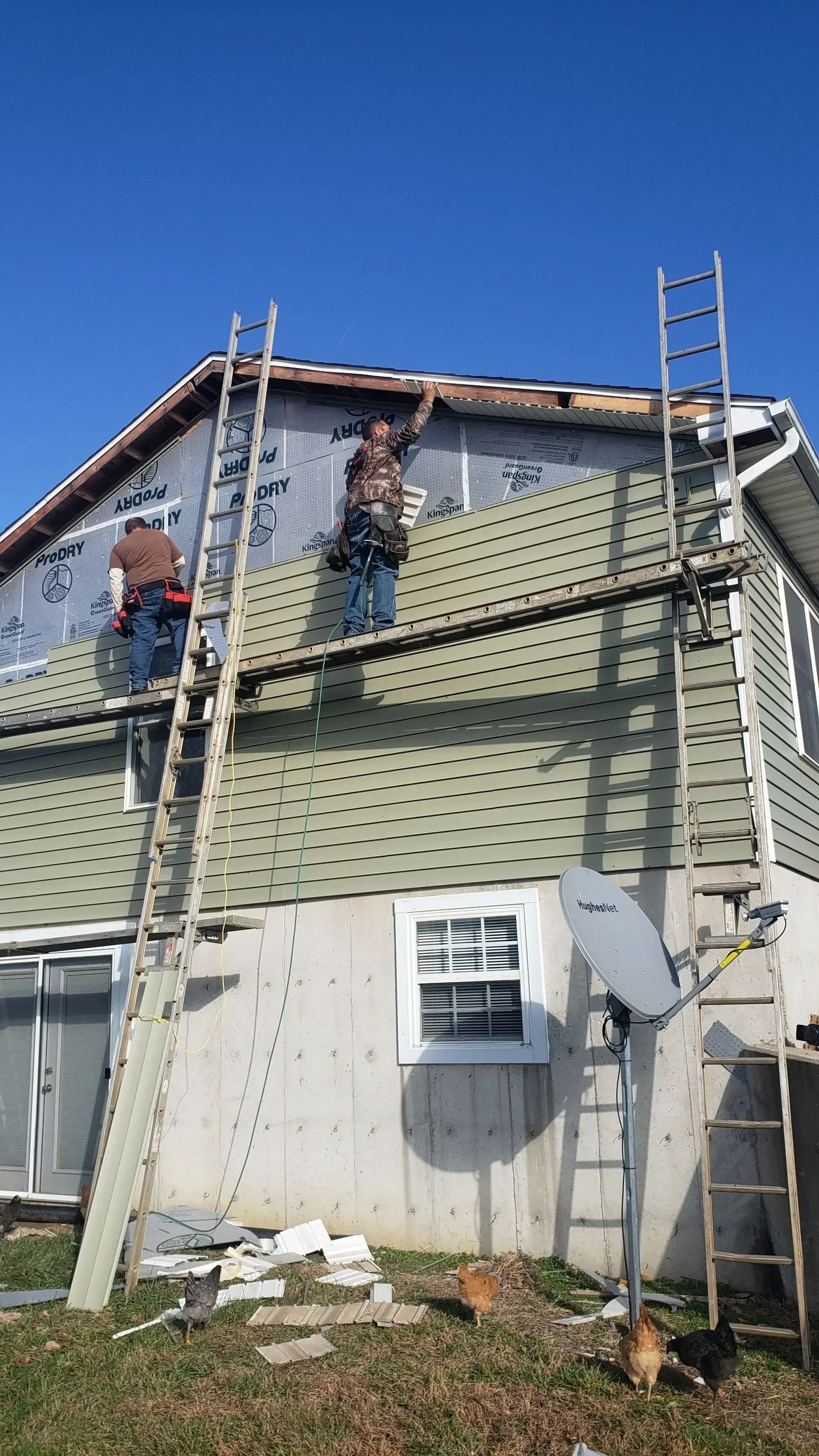 Two workers on ladders install green siding on the upper exterior wall of a two-story home.