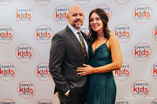 A man in a suit and a woman in a teal dress pose smiling together in front of a branded Carney’s Kids Foundation backdrop.