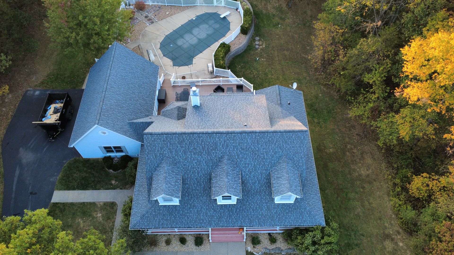 Aerial view of a house with a blue-grey roof, three dormers, a covered pool in the backyard, and a driveway with a dumpster.