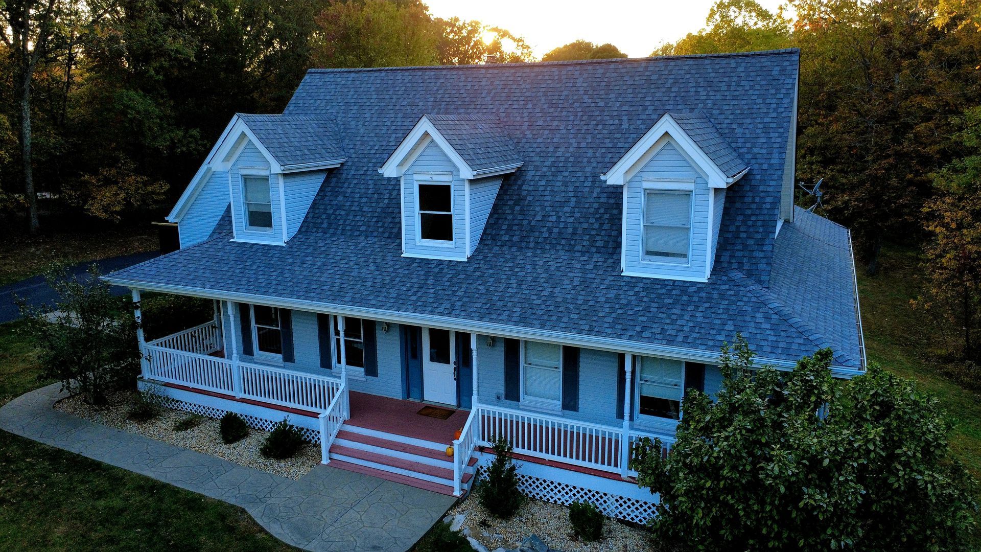 A two-story light blue house with a wrap-around porch and three dormer windows, surrounded by trees at sunset.