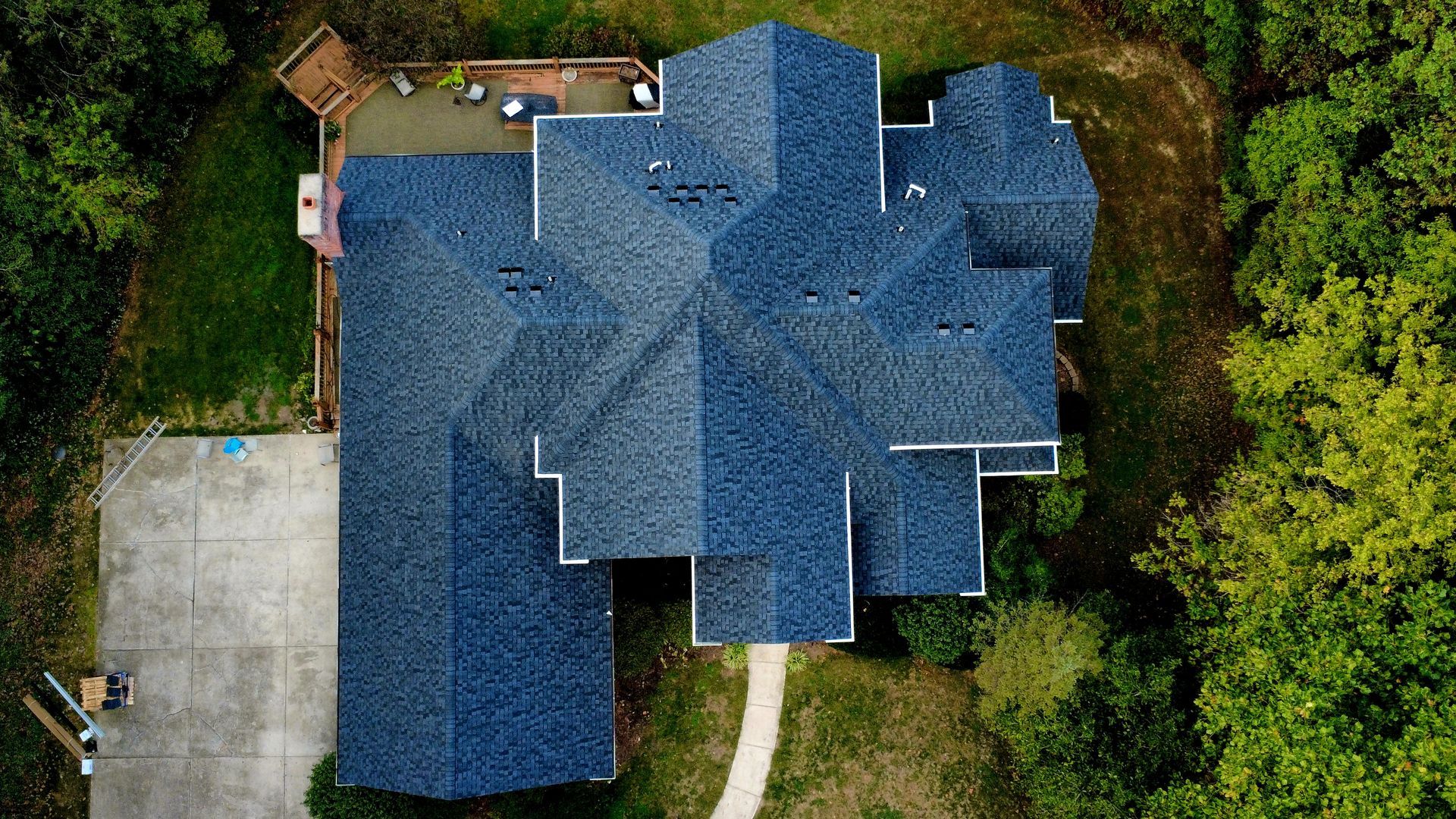 An aerial view of a blue-shingled roof on a residential house surrounded by trees and a concrete driveway.