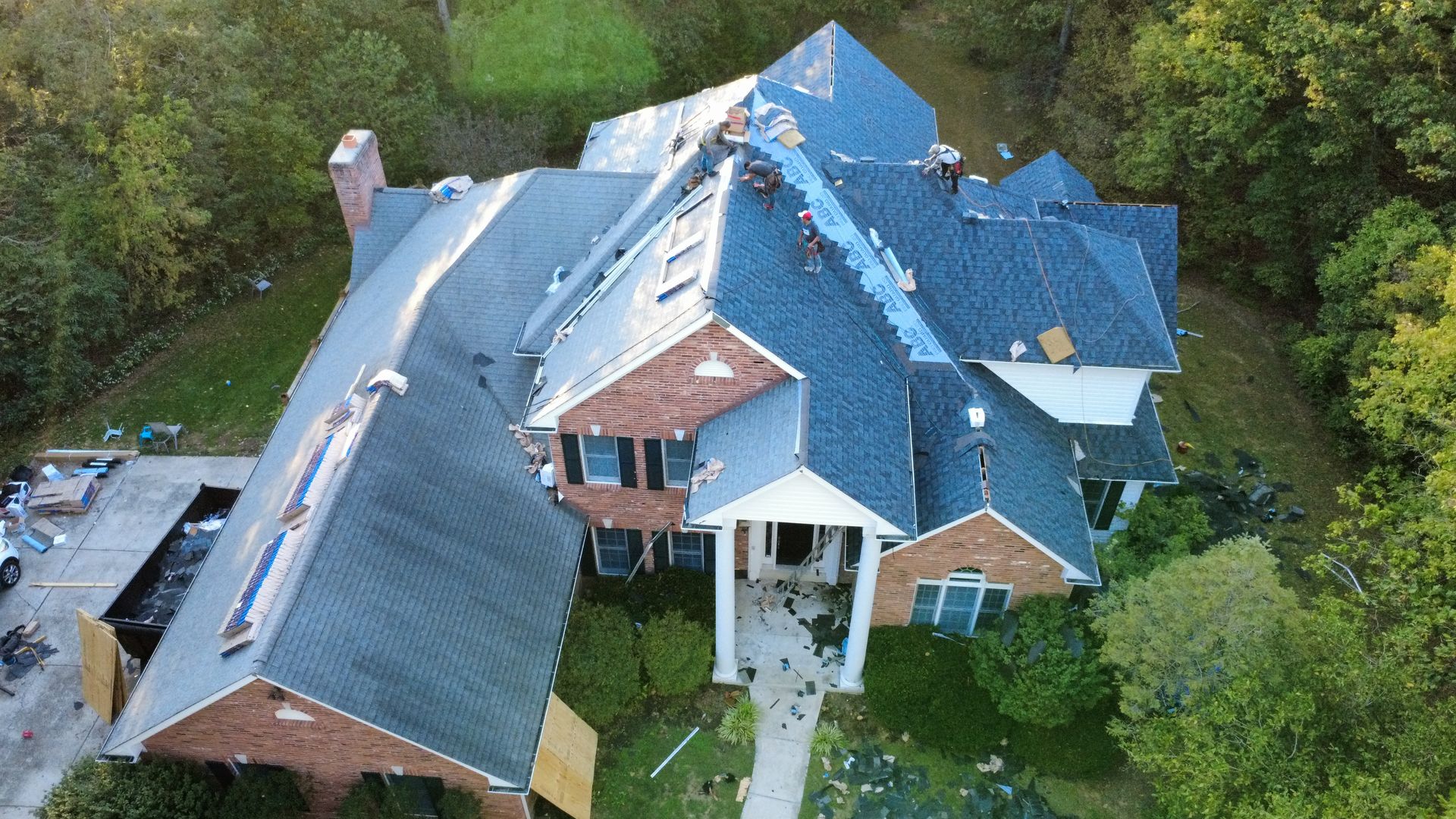 Aerial view of a large residential brick house undergoing roof repairs, with construction materials in the driveway.